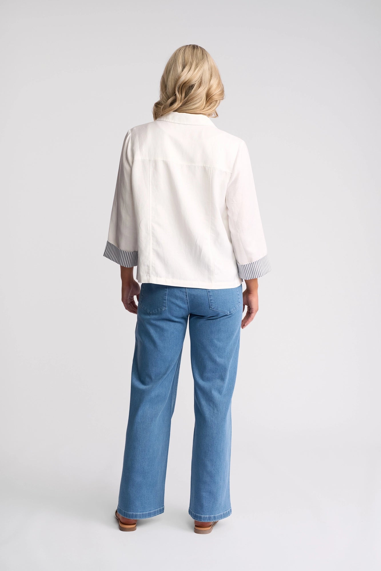 A person with blonde hair, dressed in the Vassalli Honey Jacket - White—a lightweight linen-viscose jacket featuring striped cuffs—paired with blue jeans and brown shoes, stands facing away against a plain white background.