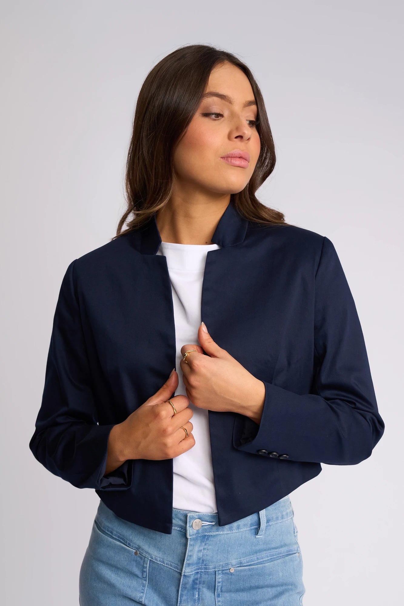 A woman models the Vassalli Midas Jacket in navy, layered over a white shirt and light blue jeans. She gazes to the side, holding the jacket open with one hand against a plain light gray background.