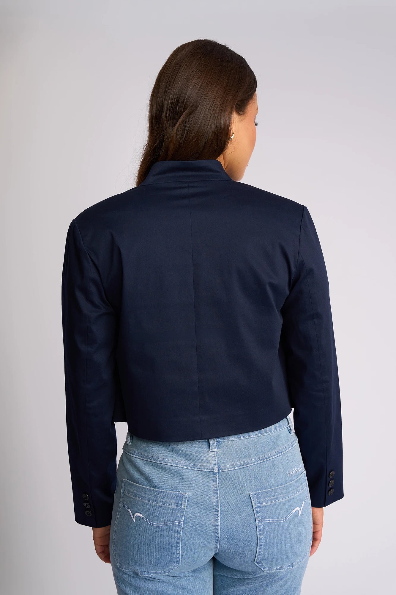 A woman is standing with her back to the camera, wearing the Vassalli Midas Jacket - Navy and light blue jeans. The background is plain and light-colored.