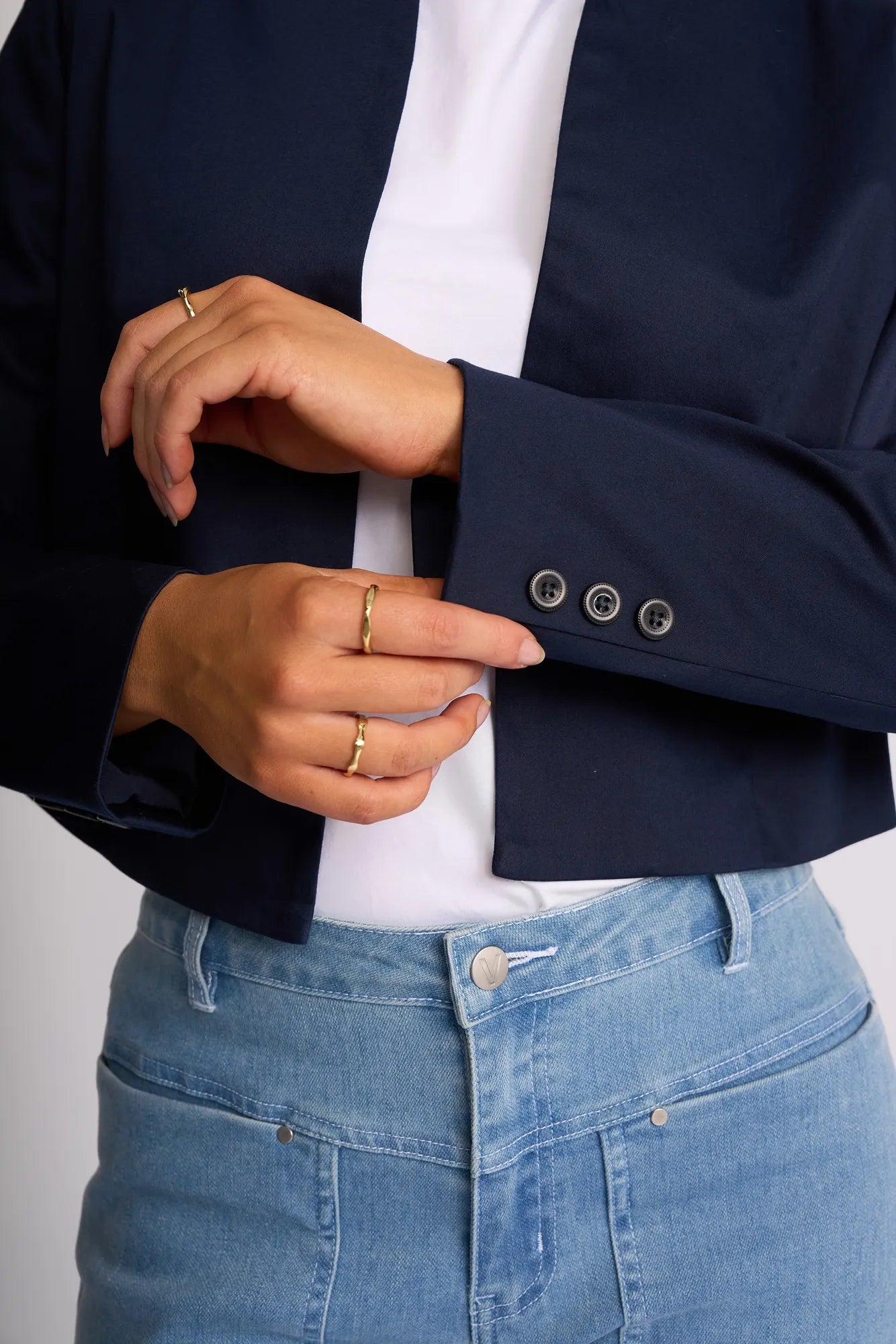 A person wearing the Vassalli Midas Jacket in navy over a white shirt and light blue jeans, adjusting the jacket’s cuff with one hand. Several gold rings are visible on their fingers.