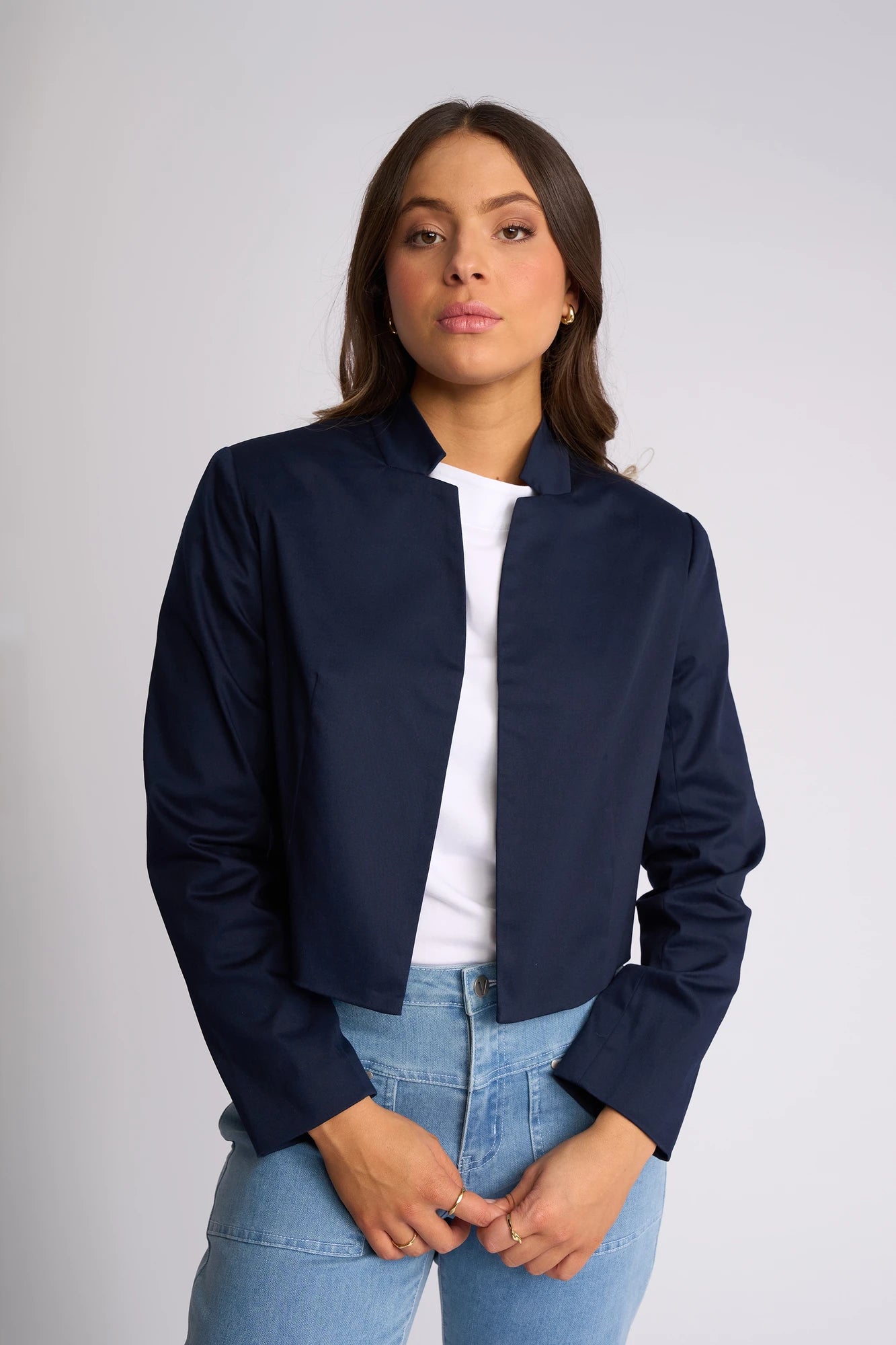 A woman with long brown hair wears a stylish Vassalli Midas Jacket in Navy over a white top and light blue jeans. She stands against a plain, light-colored background, looking directly at the camera with a neutral expression.