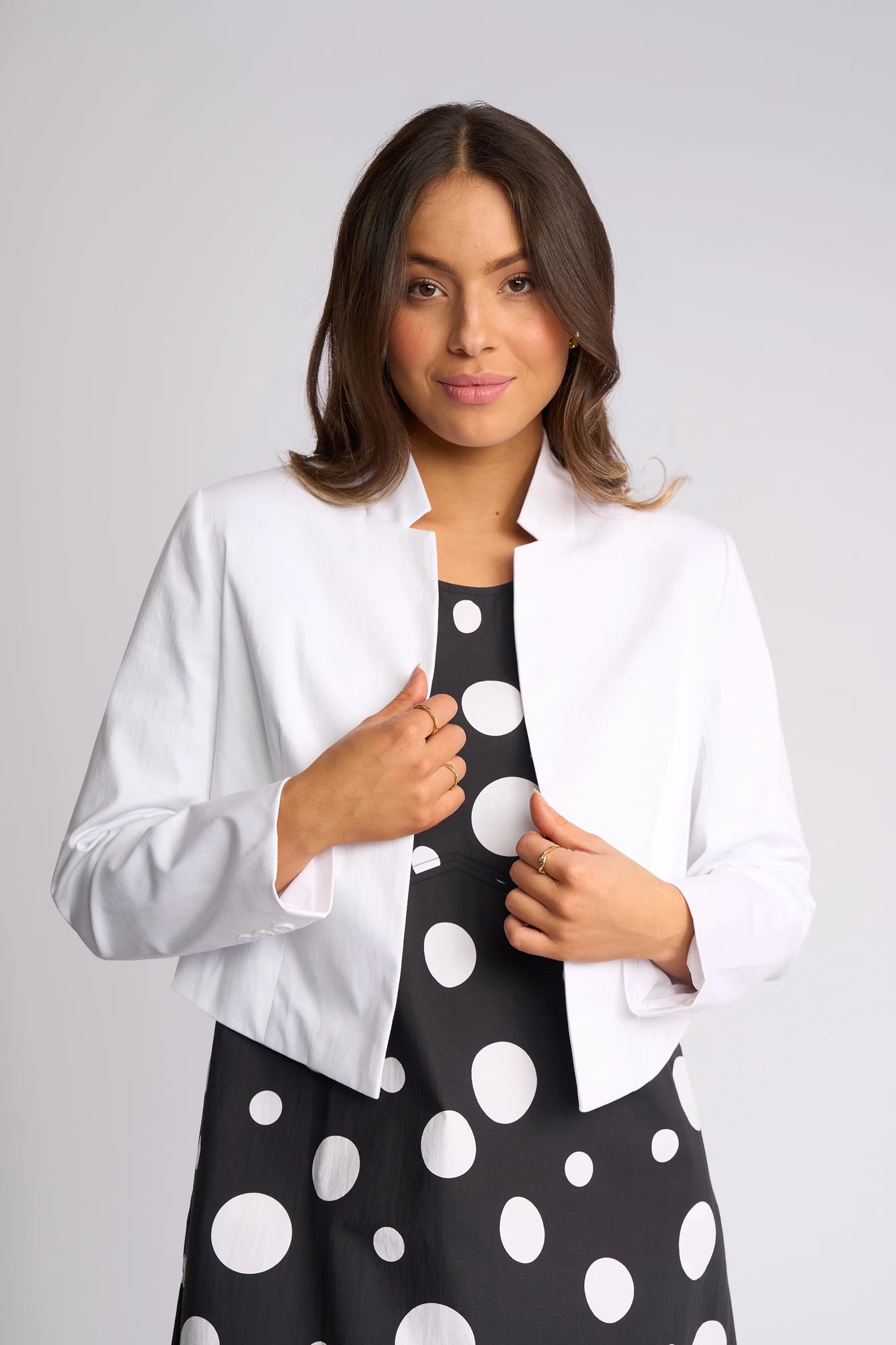 A woman with long brown hair wears the Vassalli Midas Jacket in white over a black dress with large white polka dots, standing against a light gray background and looking at the camera with a slight smile.