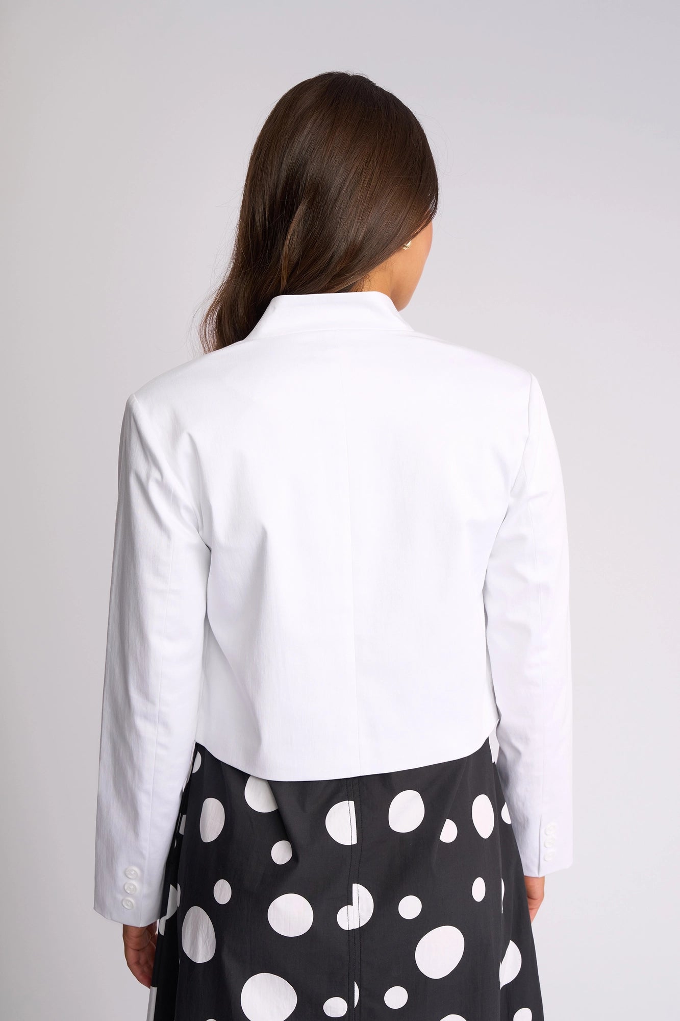 A person with long brown hair, seen from behind, wears the Vassalli Midas Jacket in white paired with a black skirt featuring large white polka dots, standing against a plain light background.