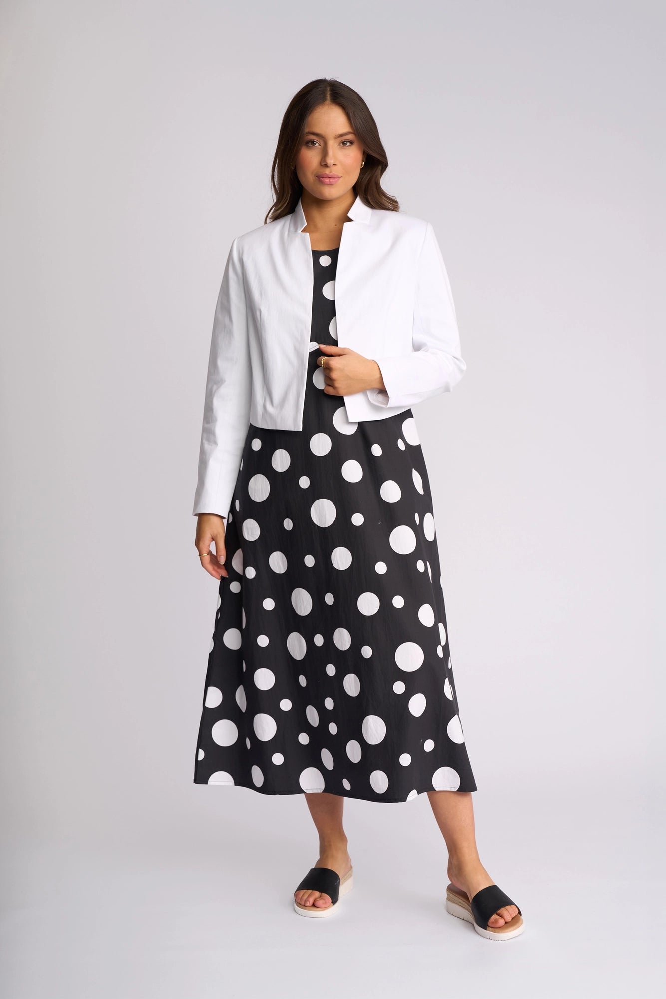A woman models the Vassalli Midas Jacket in white, layered over a black dress with bold white polka dots and styled with black open-toe sandals, set against a plain light background.