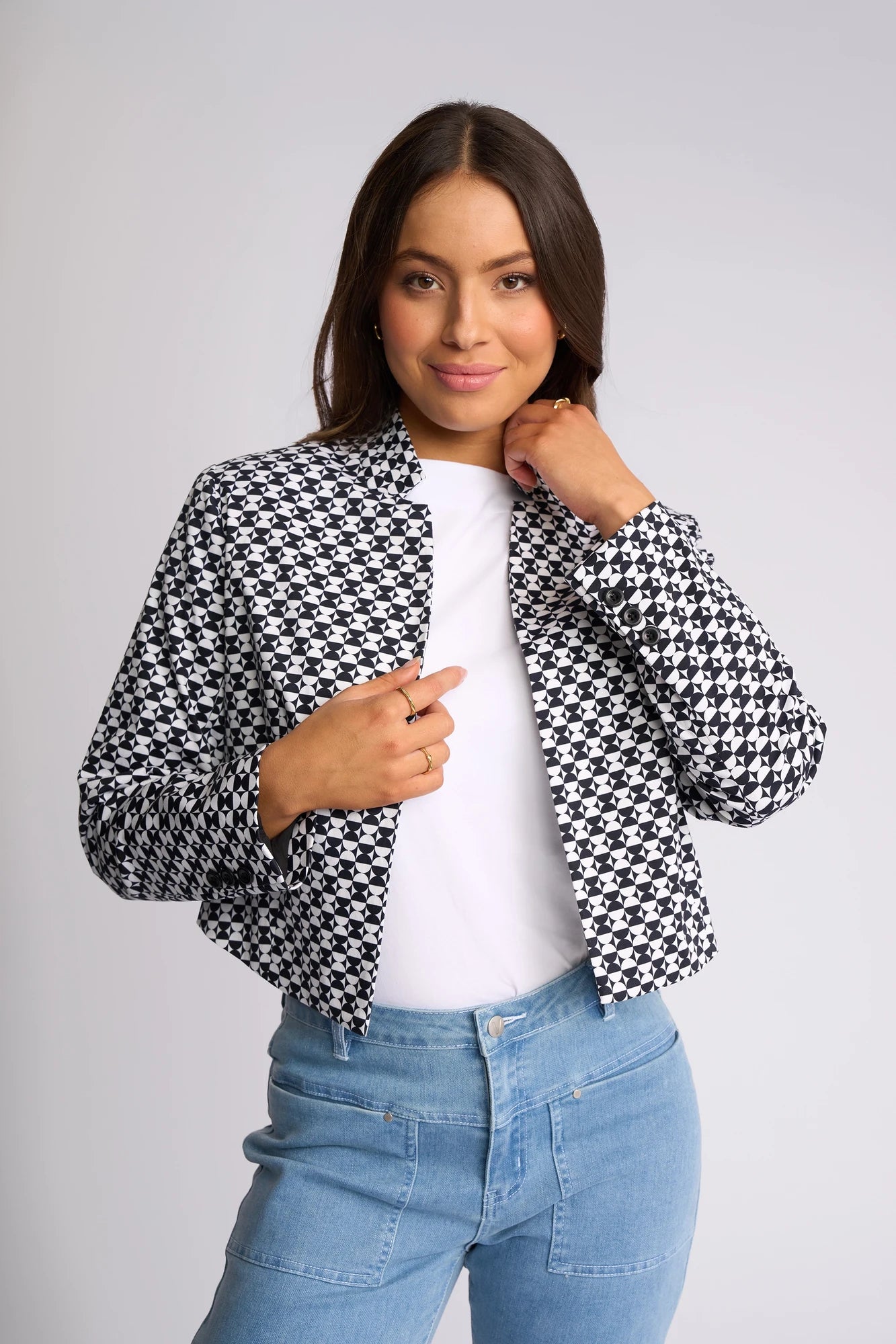 A woman stands against a light background, smiling with one hand touching her collar, wearing the Vassalli Midas Jacket - Dixie—a black-and-white houndstooth design—layered over a white shirt and paired with light blue jeans.