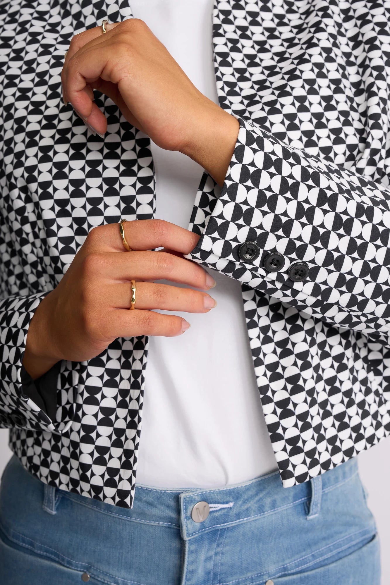 A person wearing the Vassalli Midas Jacket - Dixie, featuring a black and white geometric pattern and a collarless design, pairs it with a white shirt and light blue jeans while adjusting their sleeve. Gold rings adorn their fingers.
