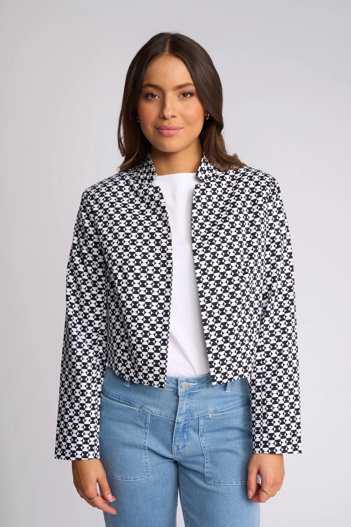 A woman stands facing the camera against a plain background, wearing the Vassalli Midas Jacket - Dixie—a collarless, black-and-white checkered jacket—layered over a white shirt and light blue jeans.