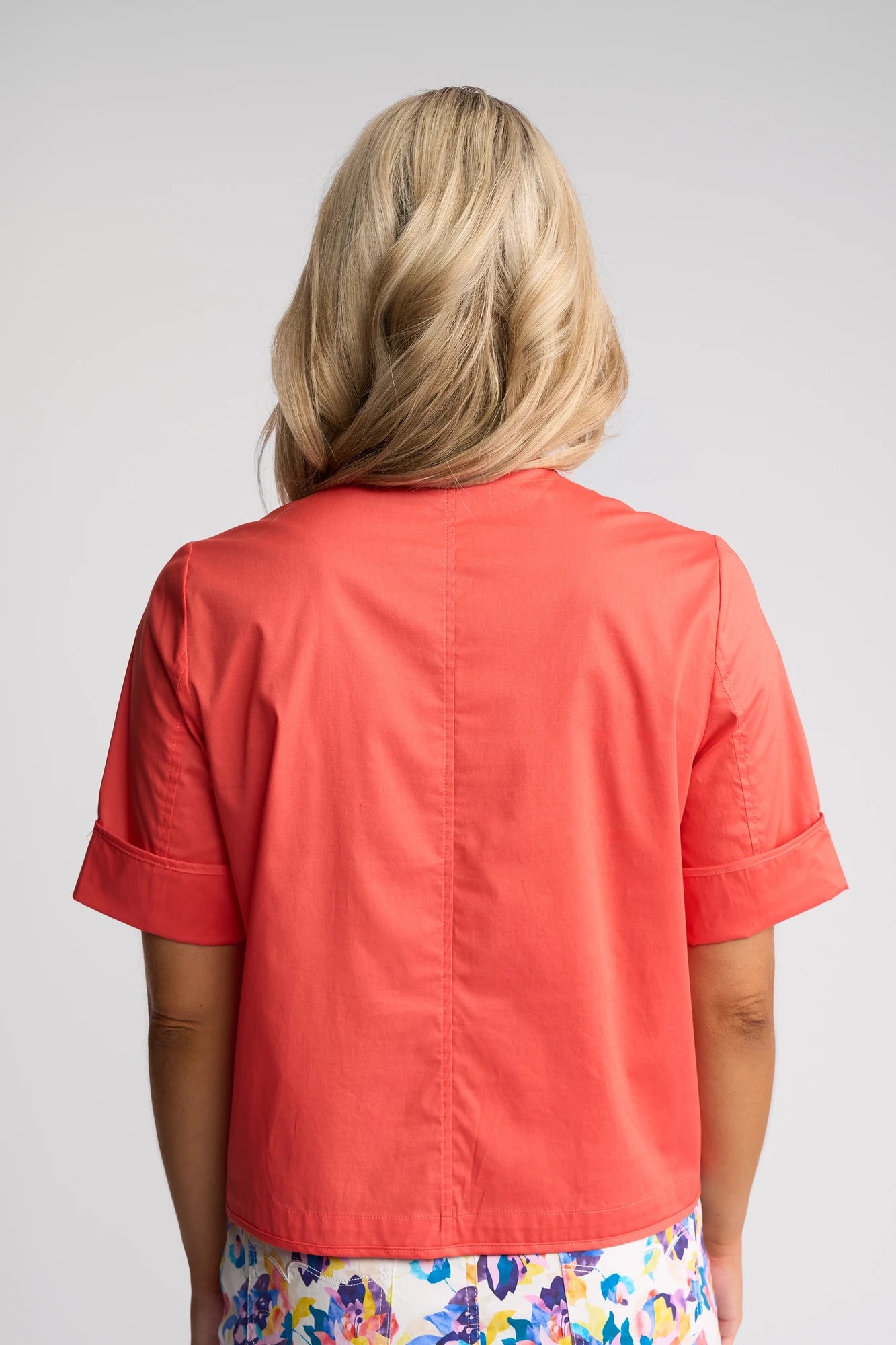 A woman with blonde hair is shown from the back, wearing a coral short-sleeve top, a colorful floral skirt, and the Vassalli Zen Jacket in Blood Orange.