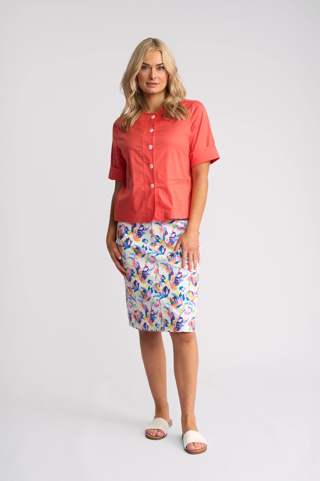 A woman stands wearing a coral button-up shirt, a colorful floral pencil skirt, and white sandals. She has shoulder-length blonde hair and is posing against a plain light background, effortlessly completing her look with the Vassalli Zen Jacket in Blood Orange—a lightweight piece crafted from stretch cotton for comfort and style.