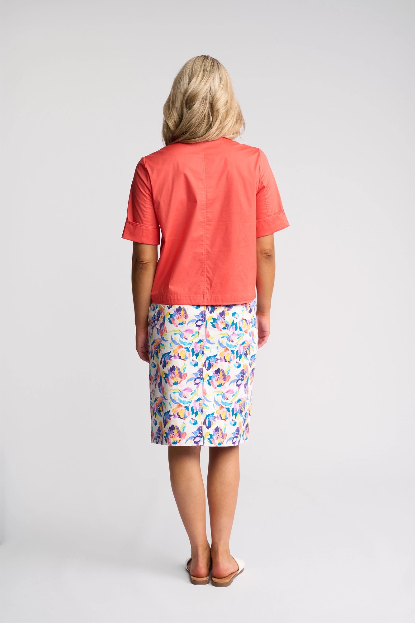 A woman stands facing away, wearing a coral short-sleeve top, a floral knee-length skirt, and white sandals, paired with the Vassalli Zen Jacket in Blood Orange stretch cotton against a plain light background.