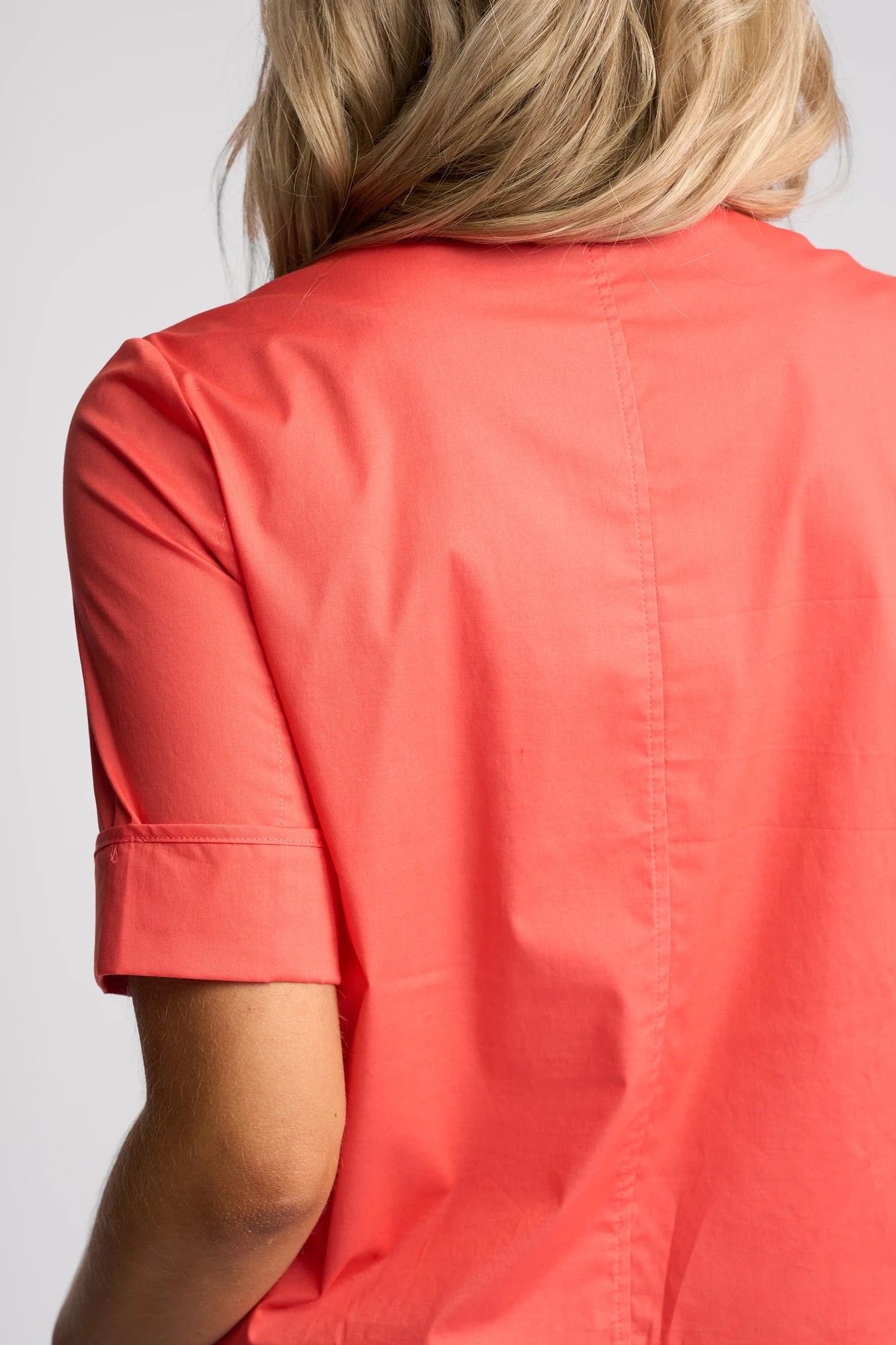 A person with blonde hair is shown from behind, wearing a short-sleeved, coral-colored shirt layered with the Vassalli Zen Jacket in Blood Orange. This lightweight stretch cotton jacket by Vassalli adds a stylish touch against the plain, light-colored background.