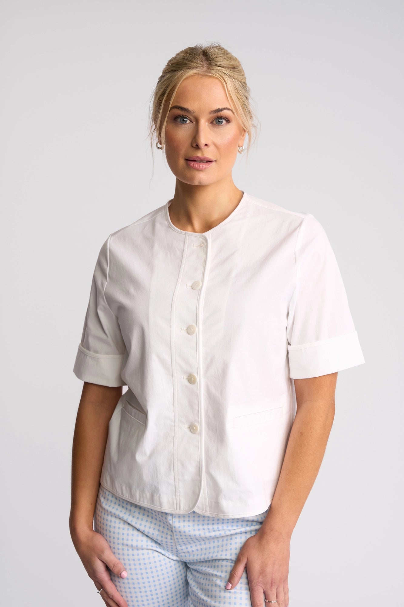 A woman with blonde hair styled in a low bun wears the Vassalli Zen Jacket in White over a white, short-sleeved button-up top and light blue checkered pants, standing against a plain, light gray background.