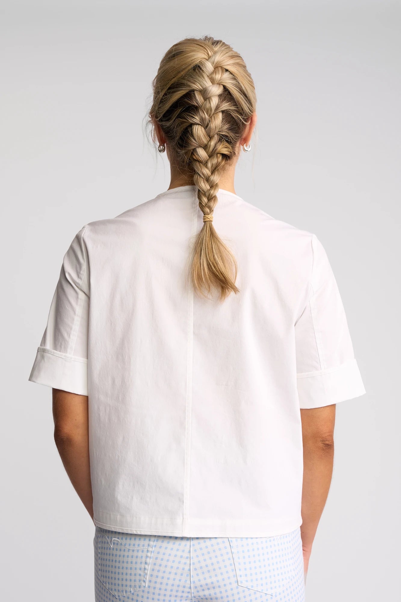 A person with blonde hair in a French braid, seen from behind against a plain background, is wearing the Vassalli Zen Jacket in white over a white short-sleeve shirt and light blue checkered pants.