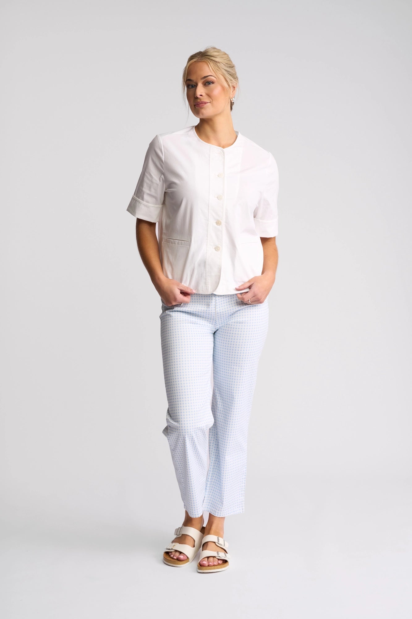 A woman stands against a plain background, wearing the Vassalli Zen Jacket in white over a white short-sleeved button-up top, paired with light blue checkered pants and white sandals. She looks to the side with her hands in her pockets.