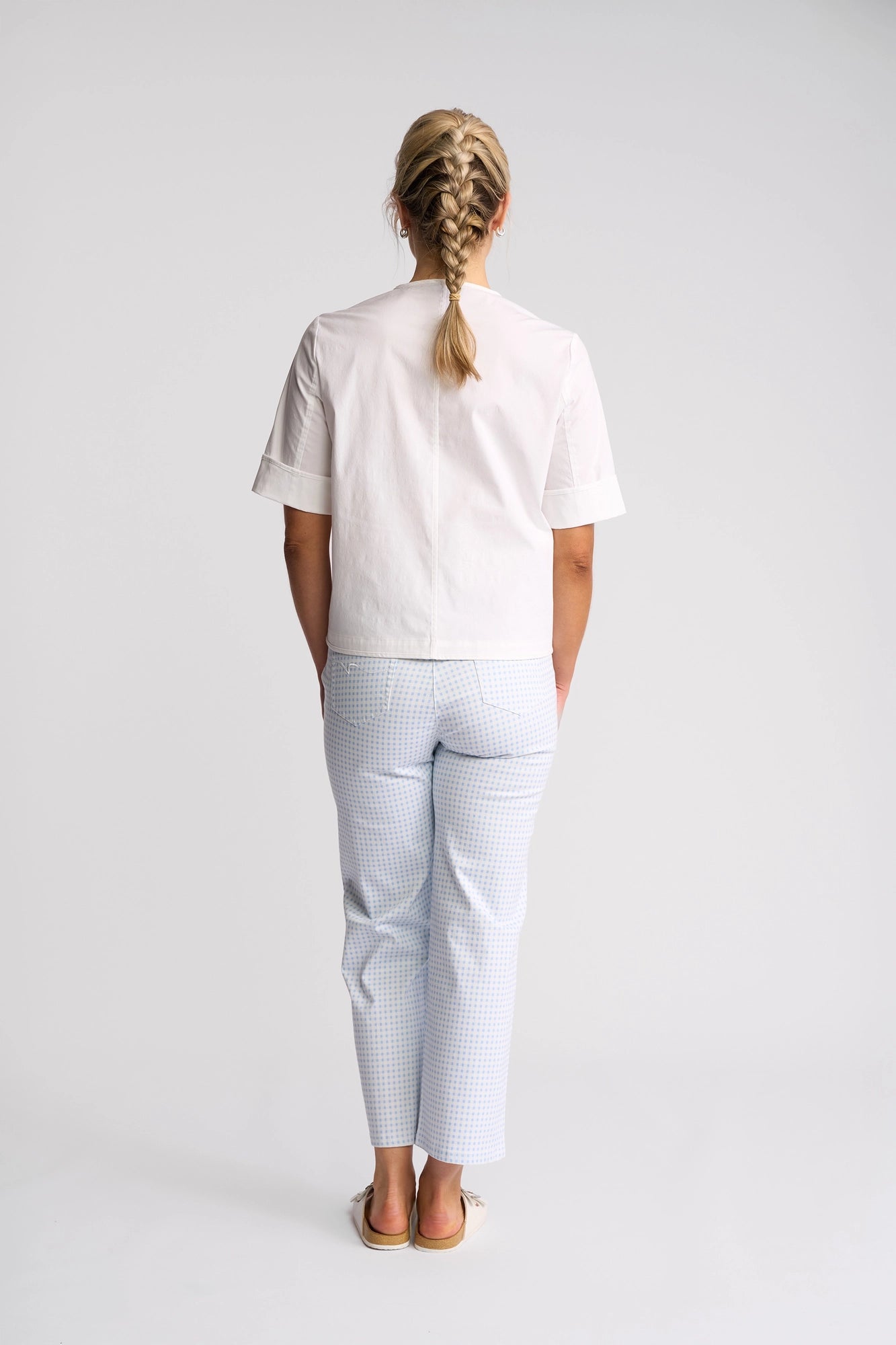 A woman with blonde hair in a braid stands facing away, wearing a white short-sleeved shirt, light blue checkered pants, and beige shoes. She has the Vassalli Zen Jacket in White draped casually over her shoulders, set against a plain white background.