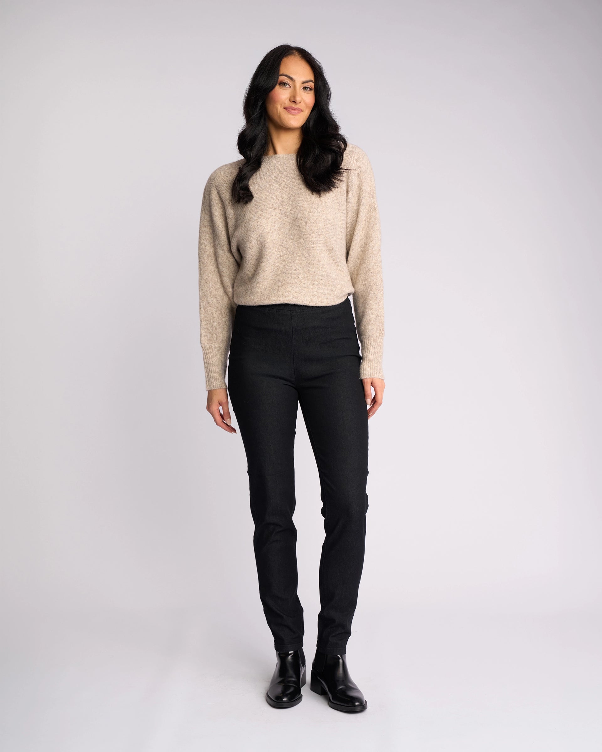 A woman with long dark hair smiles against a light background, wearing a beige sweater, black boots, and Vassalli’s Origin Pull On - Black Denim skinny-leg jeans.