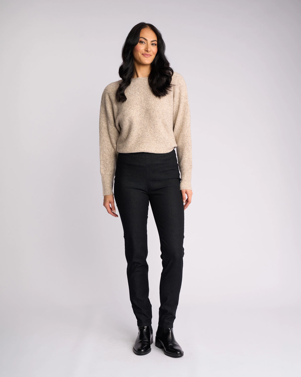 A woman with long dark hair smiles against a light background, wearing a beige sweater, black boots, and Vassalli’s Origin Pull On - Black Denim skinny-leg jeans.