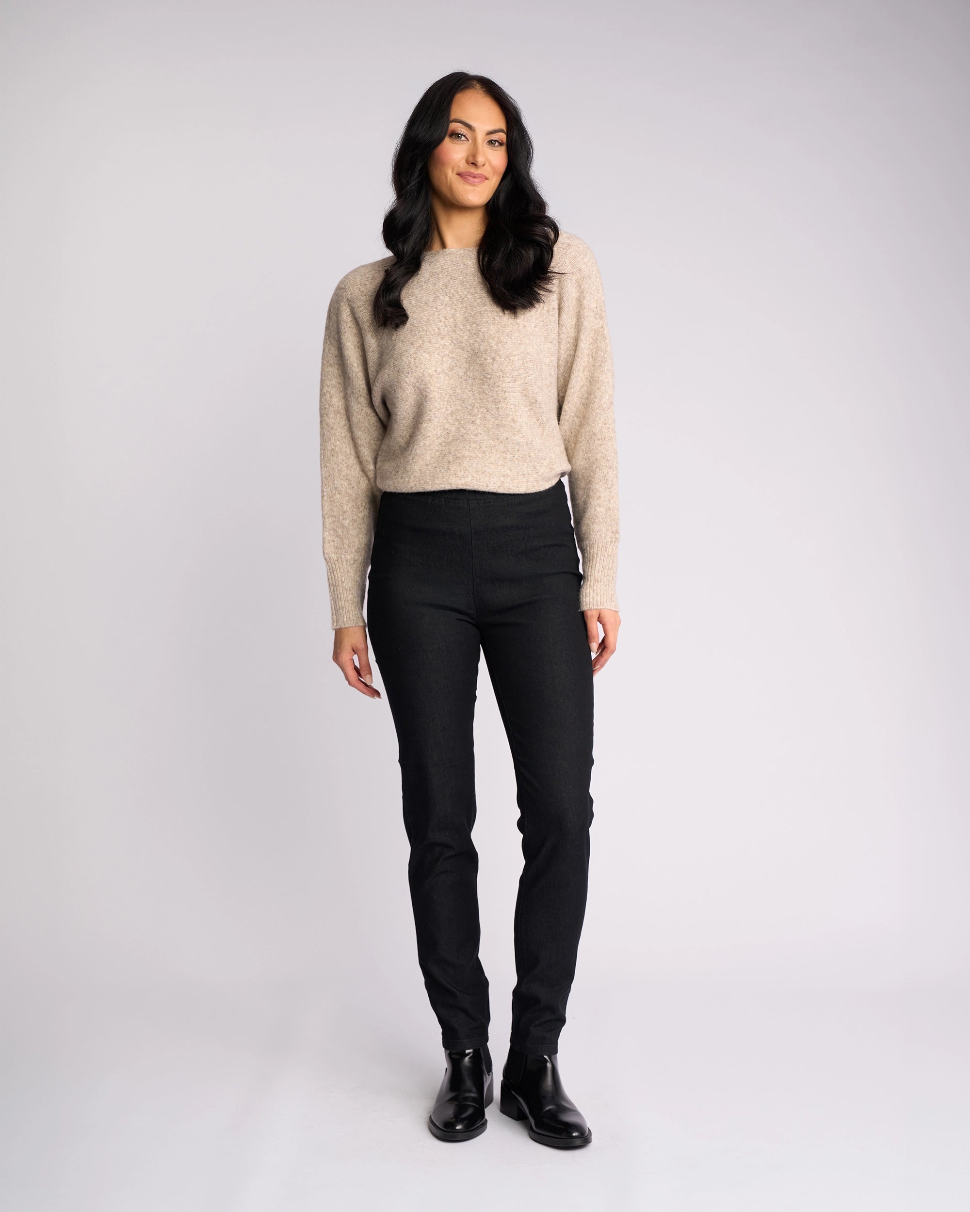 A woman with long dark hair smiles against a light background, wearing a beige sweater, black boots, and Vassalli’s Origin Pull On - Black Denim skinny-leg jeans.
