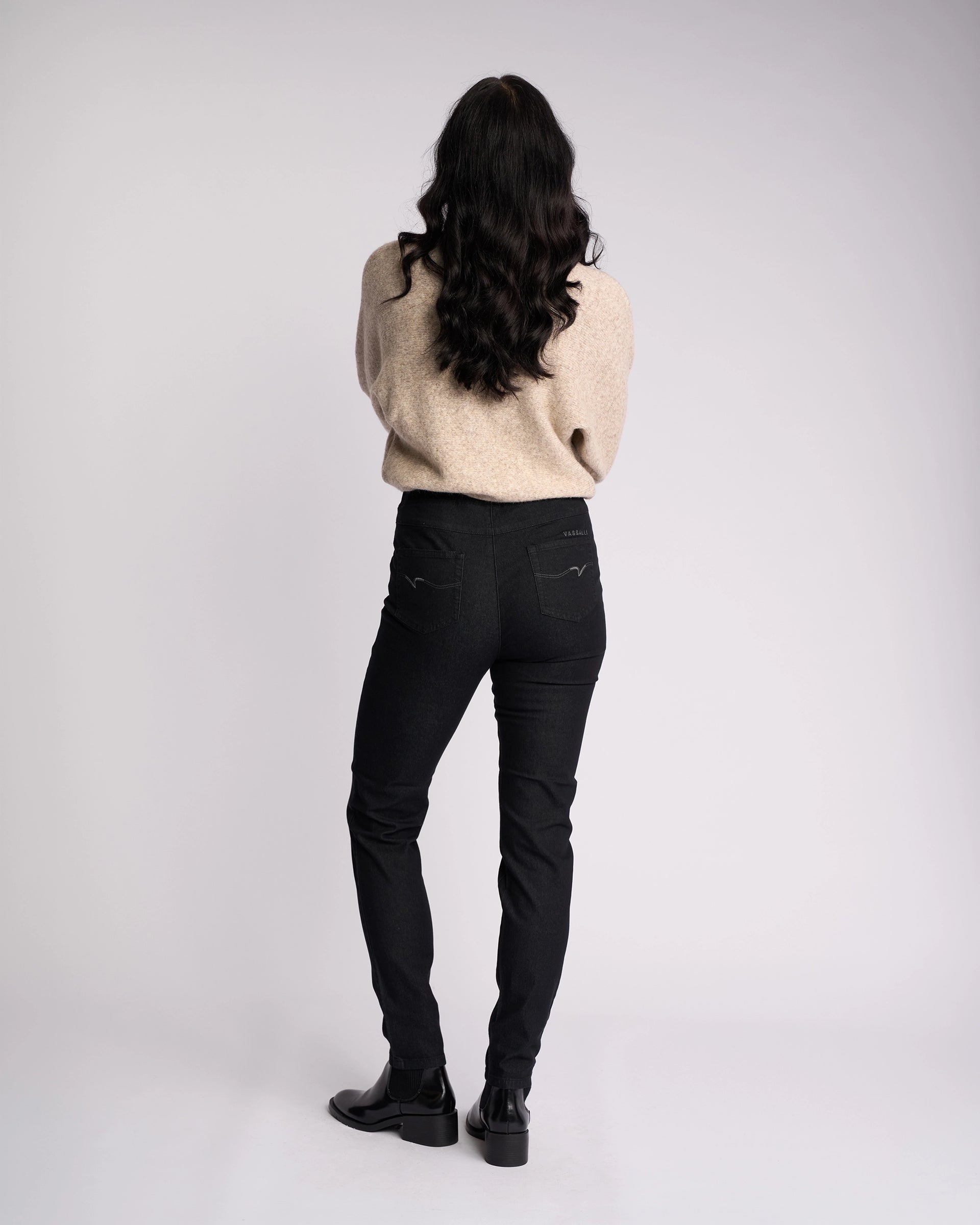 A person with long dark hair stands facing away, wearing a beige sweater and Vassalli Origin Pull On jeans in black denim, paired with black shoes against a plain, light background.