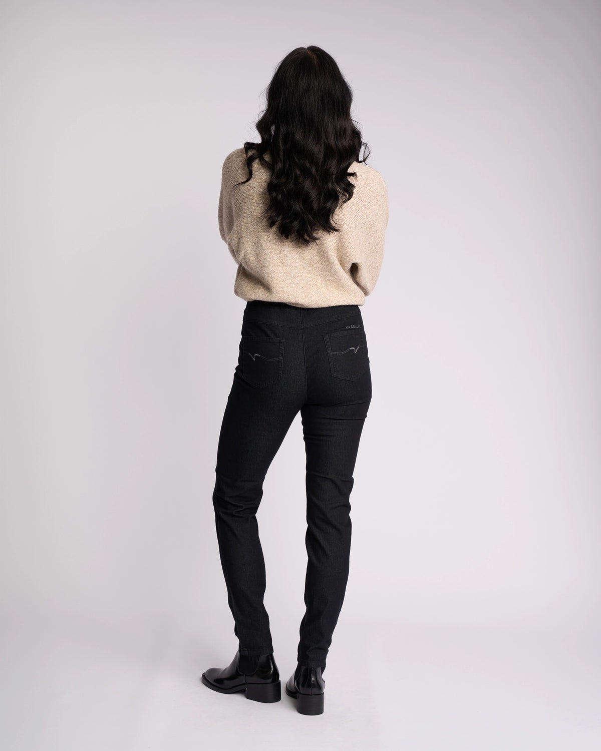 A person with long dark hair stands facing away, wearing a beige sweater and Vassalli Origin Pull On jeans in black denim, paired with black shoes against a plain, light background.
