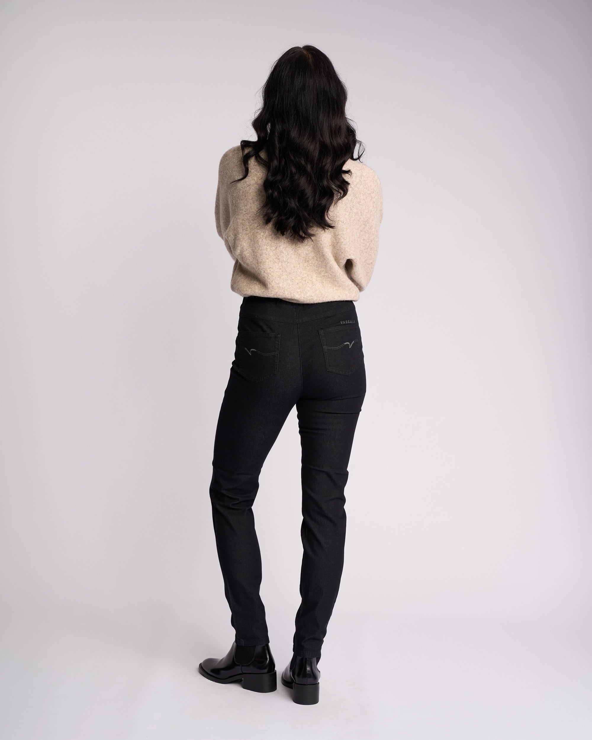 A person with long dark hair stands facing away, wearing a beige sweater and Vassalli Origin Pull On jeans in black denim, paired with black shoes against a plain, light background.