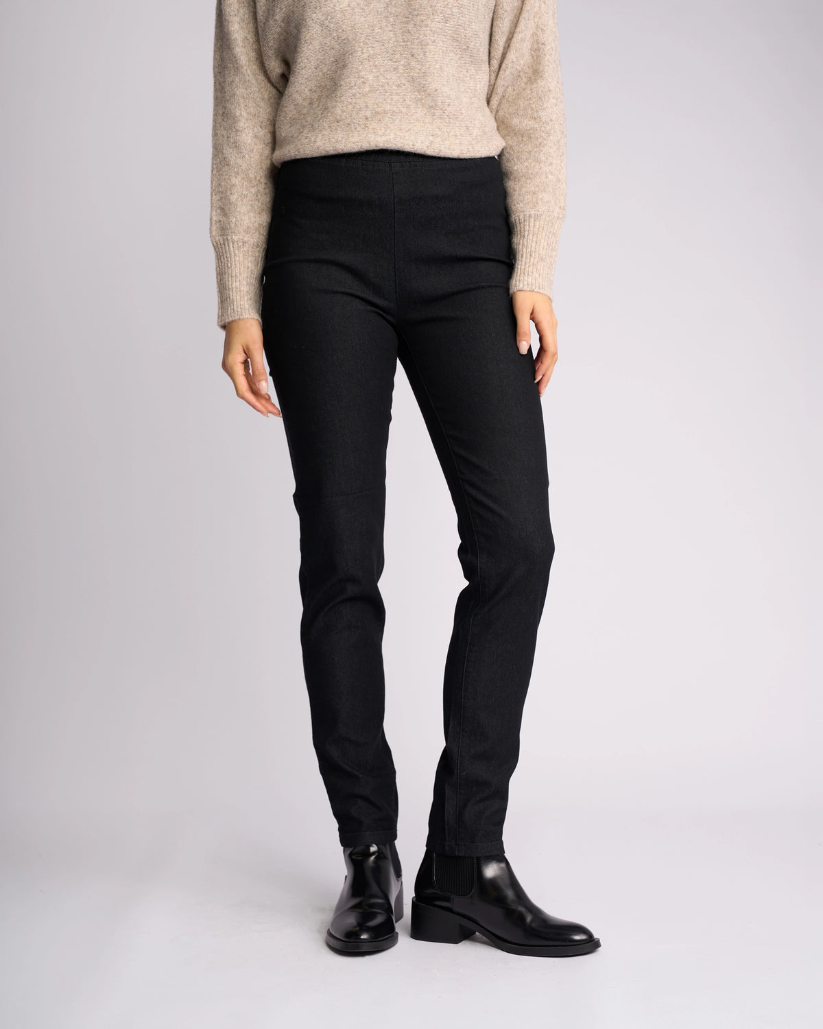 A person models the Vassalli Origin Pull On - Black Denim jeans with a beige sweater and black ankle boots, standing against a plain light background. Their arms are relaxed at their sides, and their head is not visible in the image.
