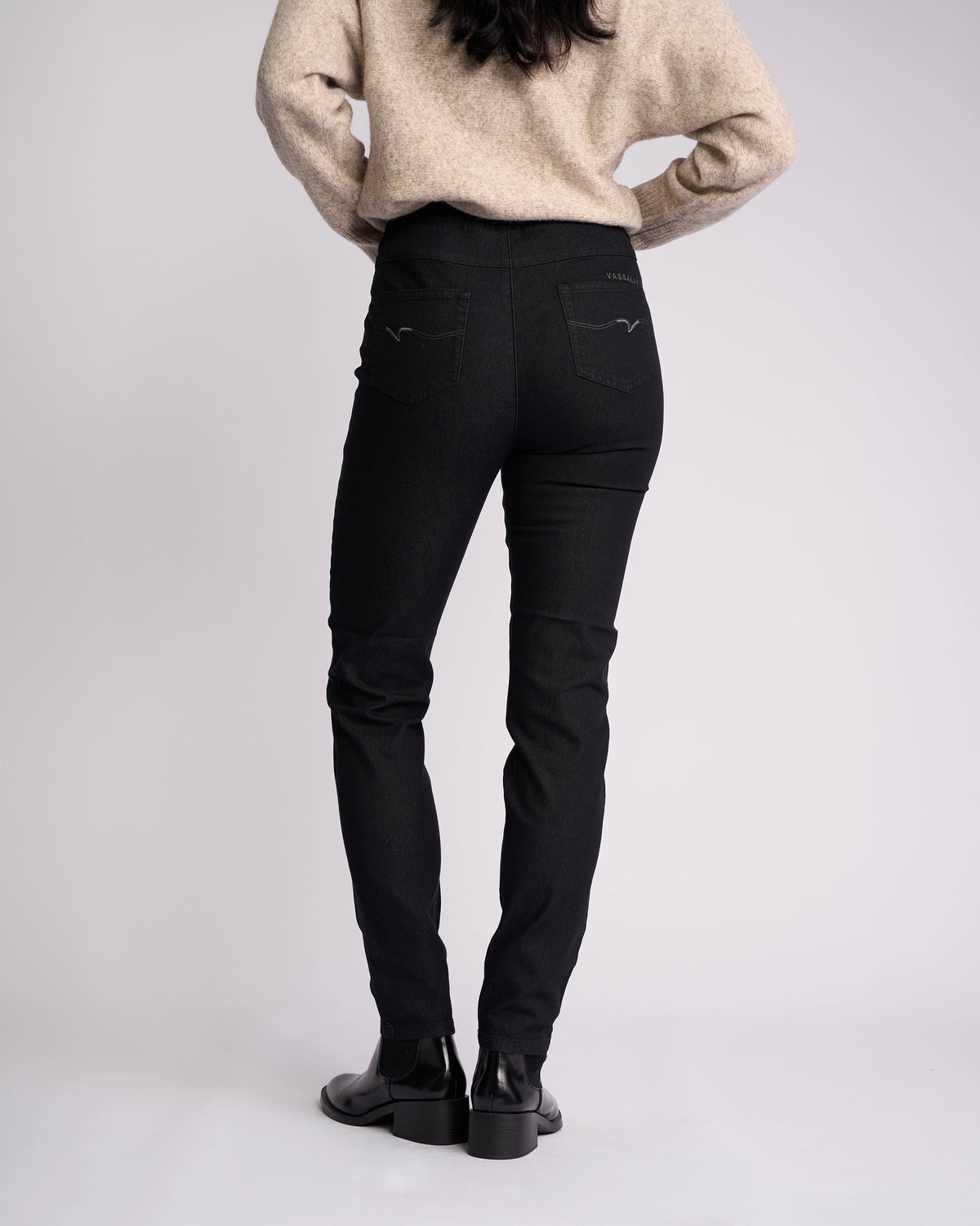 A person with long dark hair, wearing a beige sweater and Vassalli Origin Pull On - Black Denim jeans, stands with their back to the camera against a plain light background.