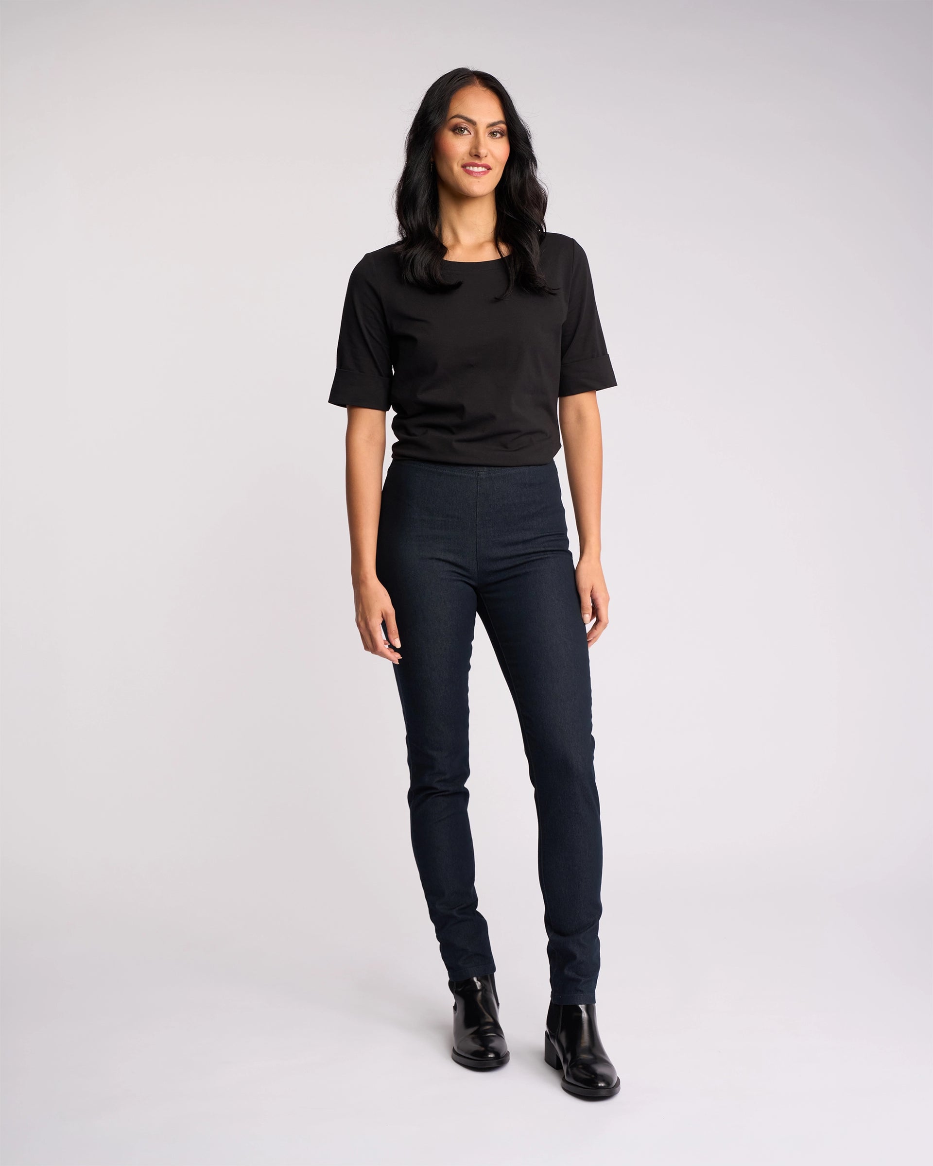 A woman with long dark hair stands against a plain light background, smiling and wearing Vassalli's Origin Pull On - Indigo Denim jeans with a black t-shirt and black ankle boots, arms relaxed at her sides.
