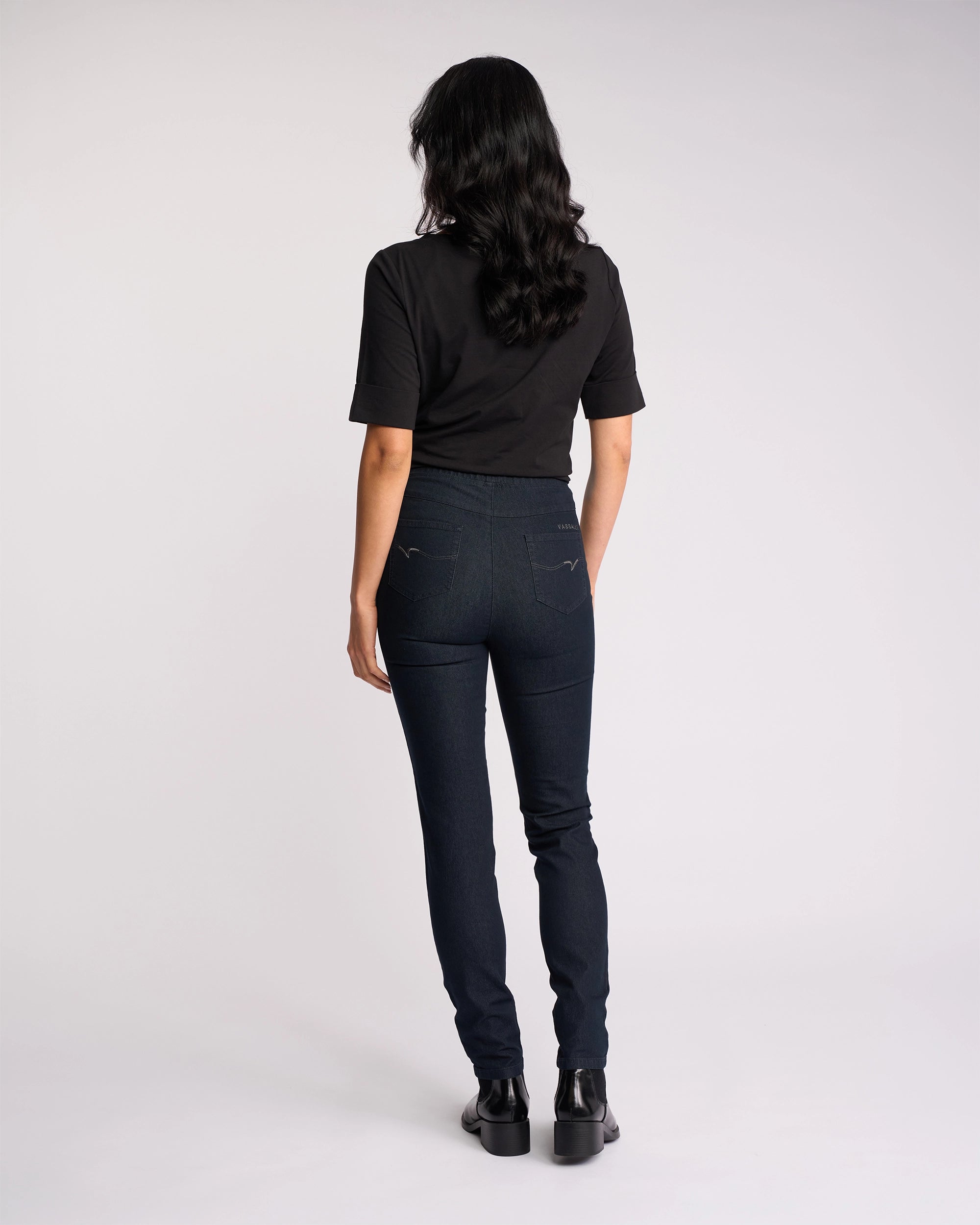 A person with long wavy hair, wearing a black t-shirt, Vassalli Origin Pull On - Indigo Denim jeans, and black shoes, stands facing away against a plain white background.