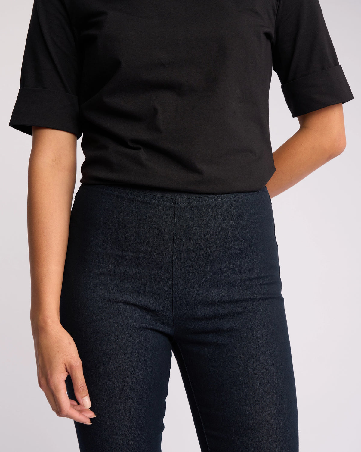 A person wearing a black short-sleeve top and Vassalli Origin Pull On Indigo Denim jeans stands against a plain light background, with one arm at their side and the other slightly bent.