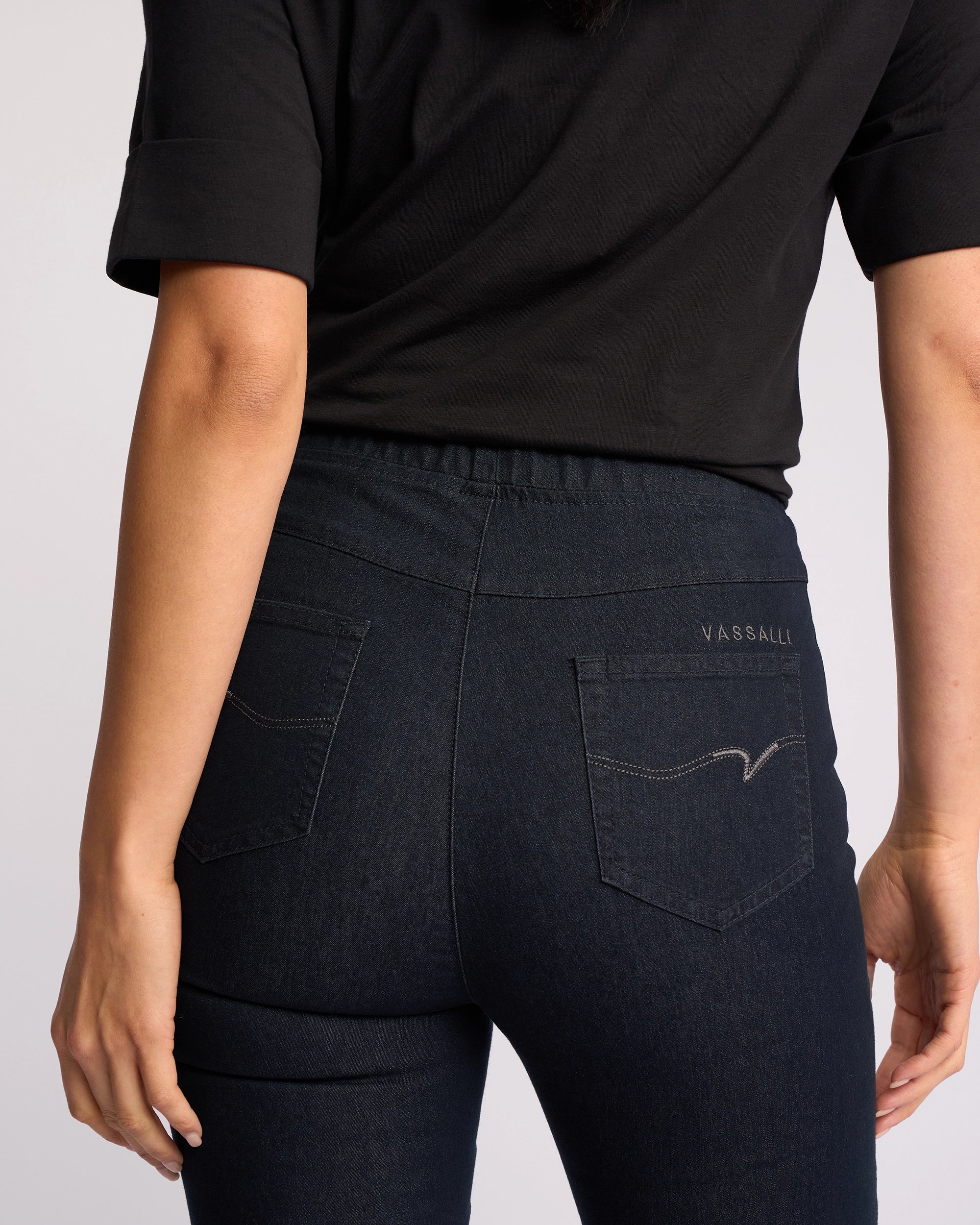 A person shown from the back waist down wears a black short-sleeve top and Vassalli Origin Pull On indigo denim jeans, featuring visible pocket stitching and Vassalli embroidery on the right pocket.