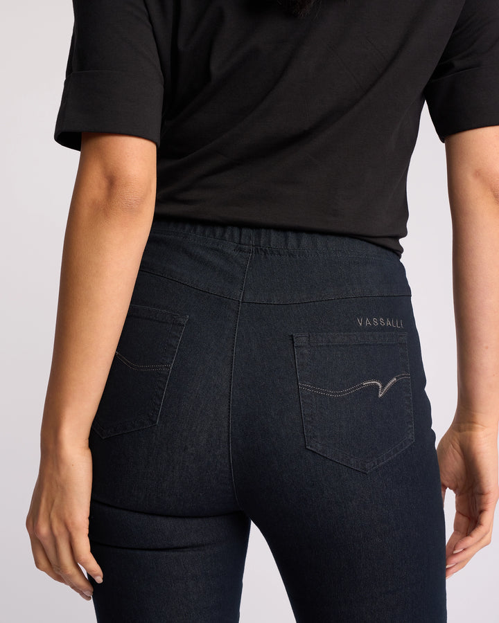 A person shown from the back waist down wears a black short-sleeve top and Vassalli Origin Pull On indigo denim jeans, featuring visible pocket stitching and Vassalli embroidery on the right pocket.
