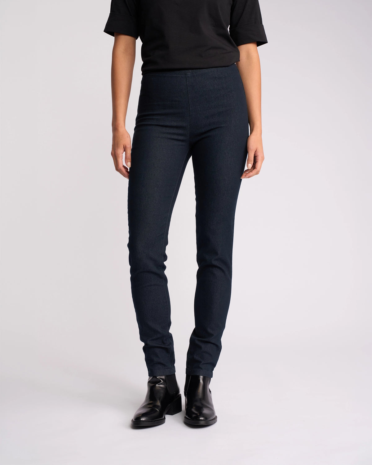 Wearing a black short-sleeve top, Vassalli Origin Pull On - Indigo Denim jeans, and black ankle boots, the person stands against a plain white background with their face not visible.