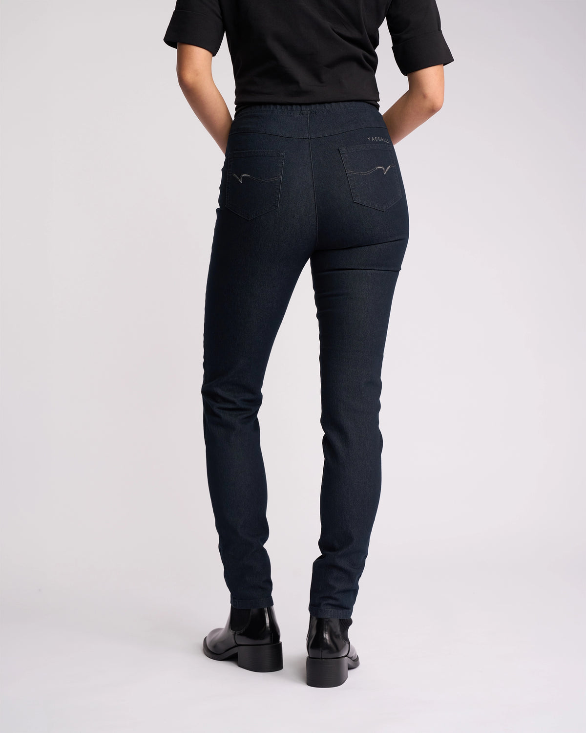 A person wearing a black short-sleeve top, Vassalli Origin Pull On - Indigo Denim jeans, and black ankle boots stands facing away from the camera against a plain light background.