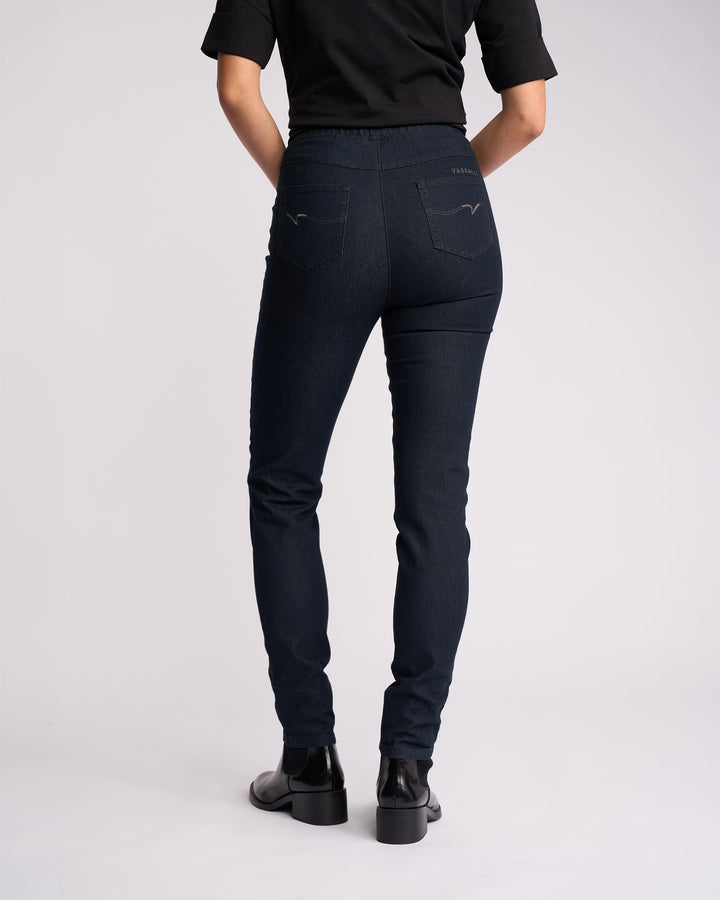 A person wearing a black short-sleeve top, Vassalli Origin Pull On - Indigo Denim jeans, and black ankle boots stands facing away from the camera against a plain light background.