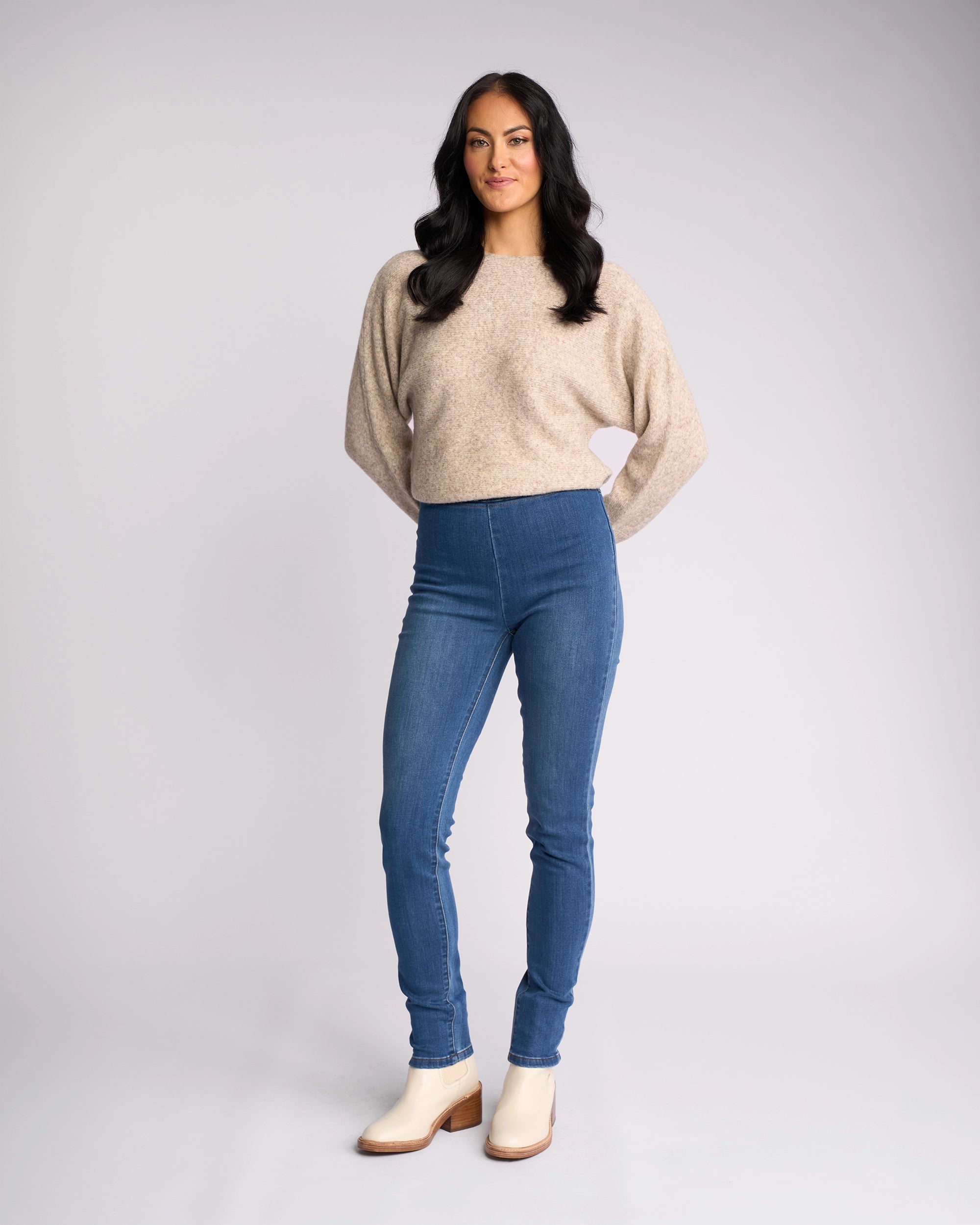 A woman with long dark hair stands facing forward, wearing a beige sweater and Vassalli Origin Pull On - New Blue Denim jeans, paired with light-colored boots, against a plain light gray background.