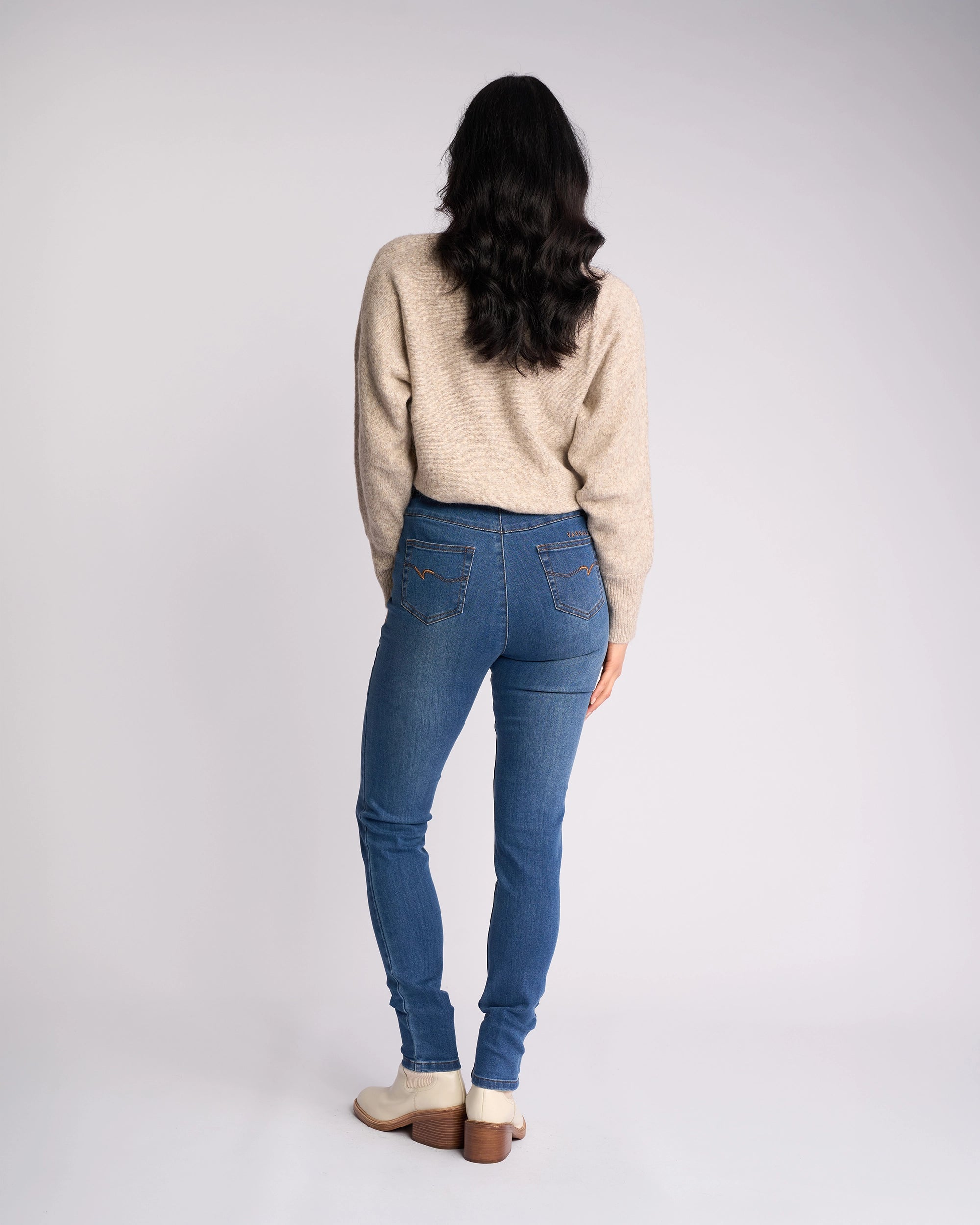 A woman with long dark hair, in a beige sweater and beige shoes, models Vassalli Origin Pull On - New Blue Denim jeans, standing facing away from the camera against a plain light background.