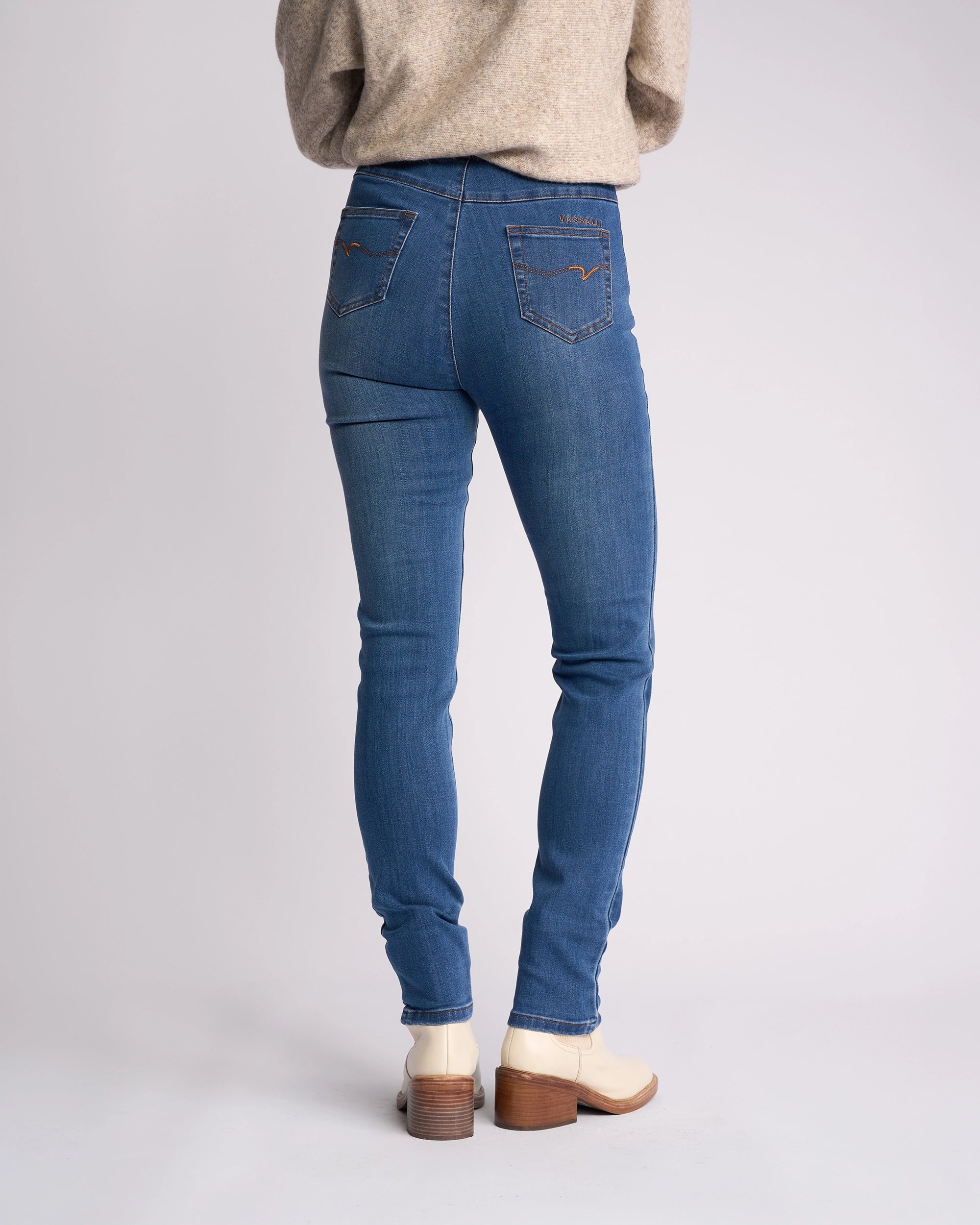 A person in a beige sweater, Vassalli Origin Pull On - New Blue Denim jeans, and cream heeled boots stands with their back to the camera against a plain light background.