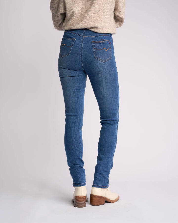 A person in a beige sweater, Vassalli Origin Pull On - New Blue Denim jeans, and cream heeled boots stands with their back to the camera against a plain light background.