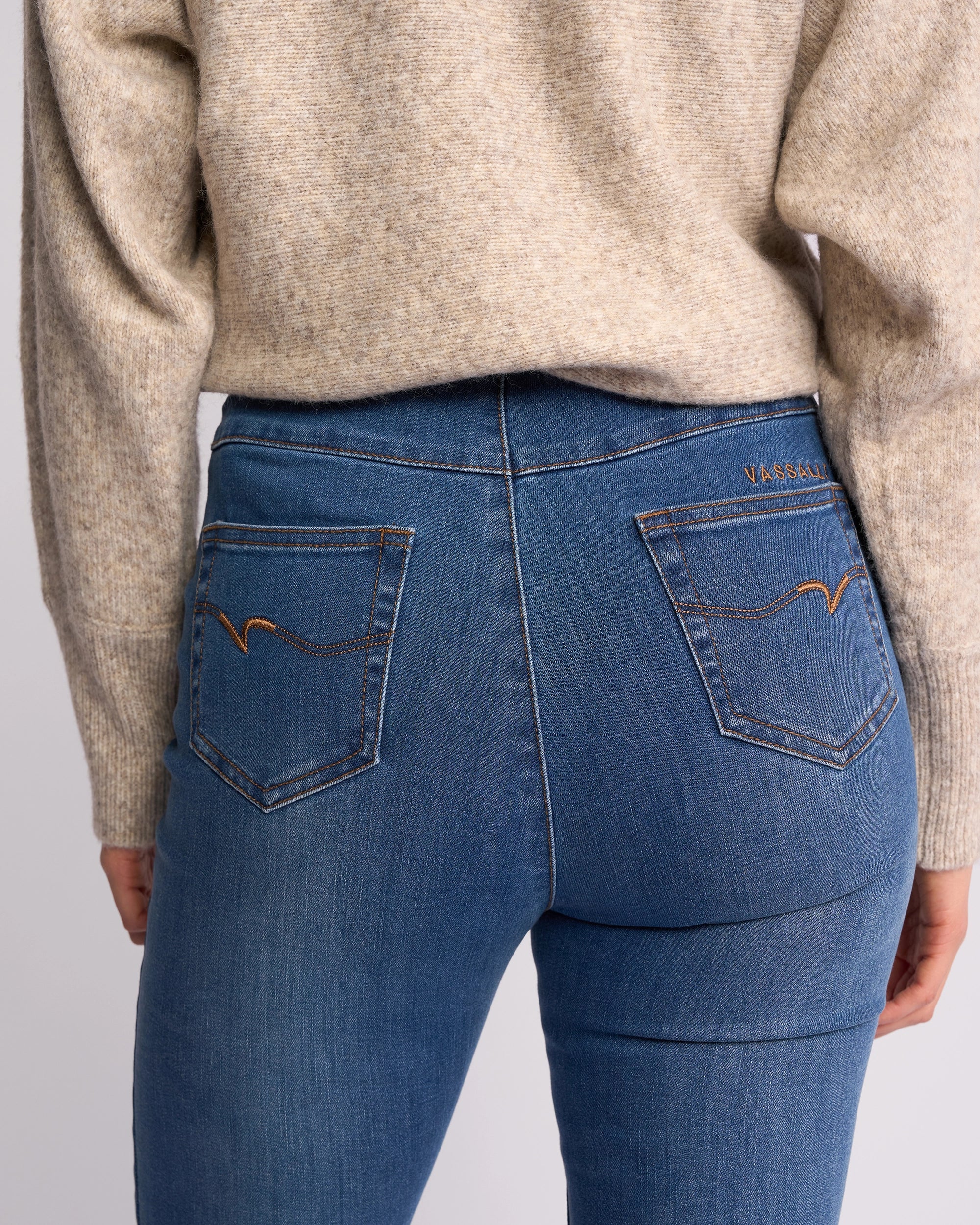 A person wearing a beige sweater and Vassalli Origin Pull On - New Blue Denim jeans is shown from the back. The high-rise jeans have two back pockets with brown stitching and a small brand label above the right pocket.