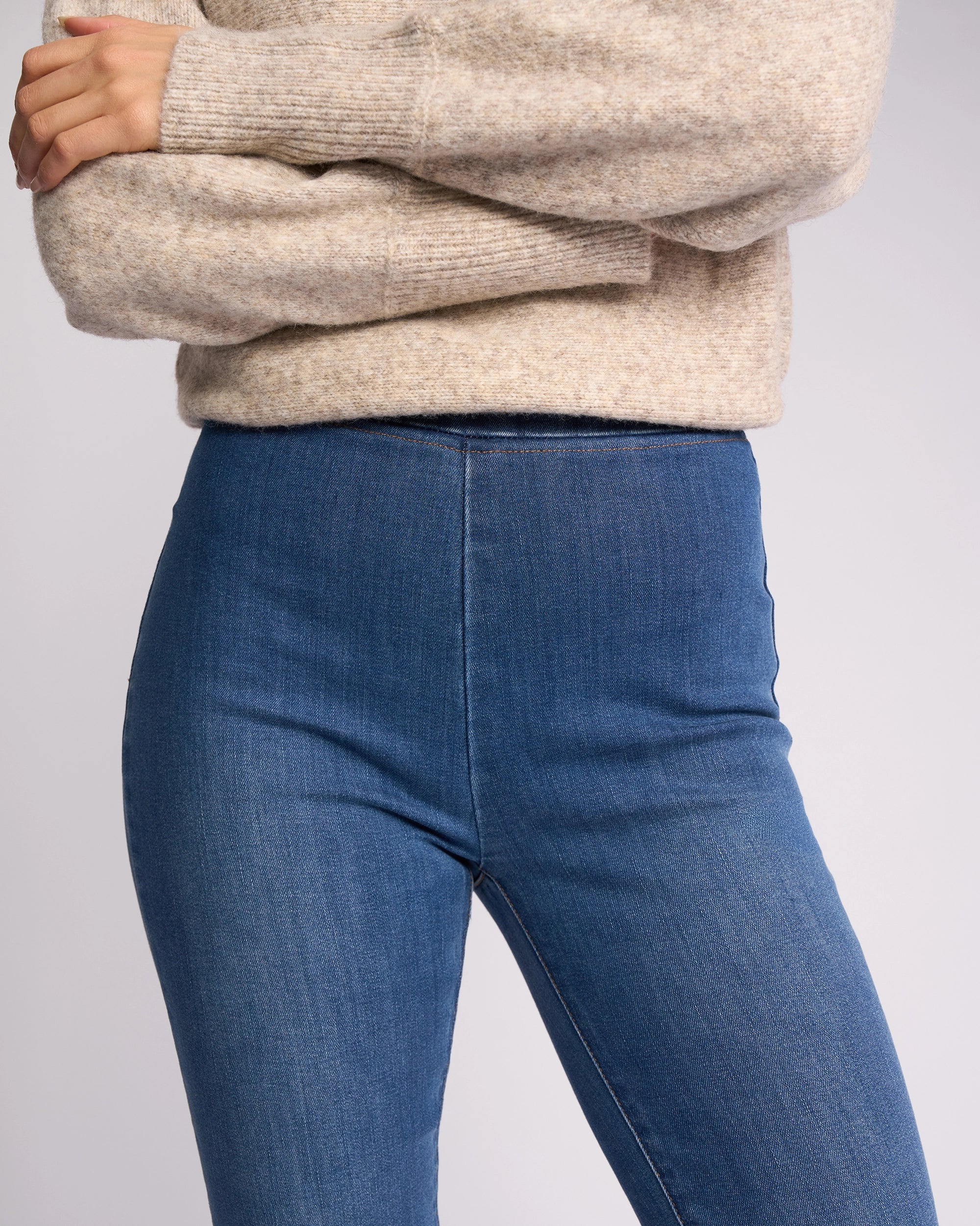 A person models Vassalli’s Origin Pull On - New Blue Denim, standing with arms crossed in a beige knit sweater, shown from shoulders to mid-thigh against a plain background.