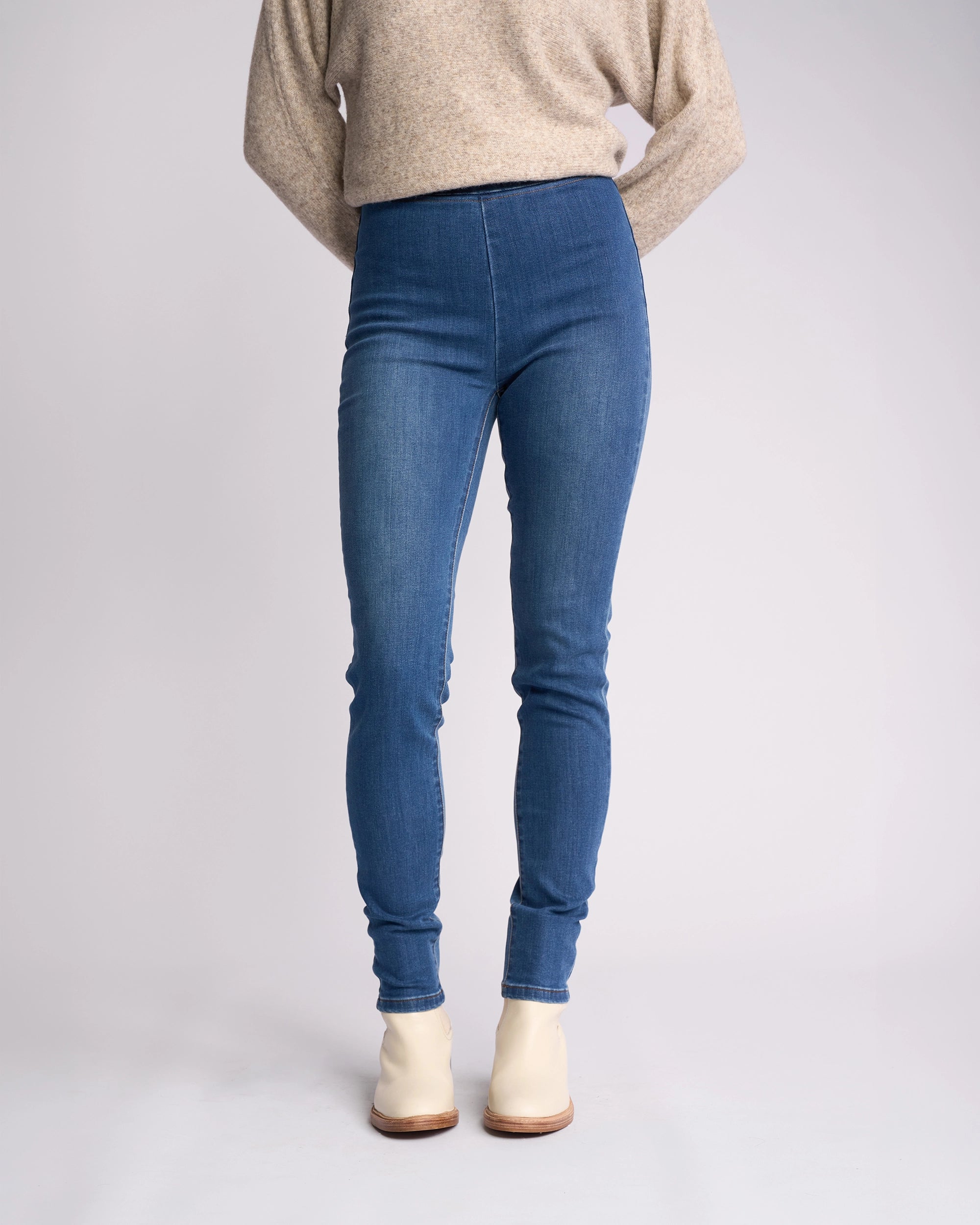 Wearing Vassalli Origin Pull On - New Blue Denim skinny jeans, a beige long-sleeve sweater, and cream ankle boots, a person stands with hands behind their back against a plain light background. Face is not visible.