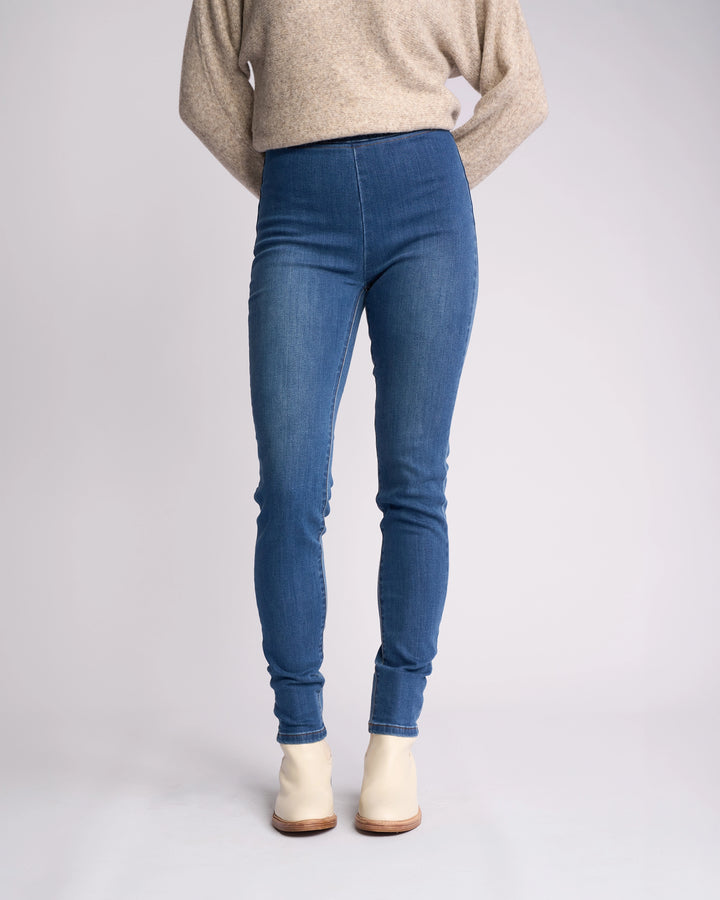 Wearing Vassalli Origin Pull On - New Blue Denim skinny jeans, a beige long-sleeve sweater, and cream ankle boots, a person stands with hands behind their back against a plain light background. Face is not visible.