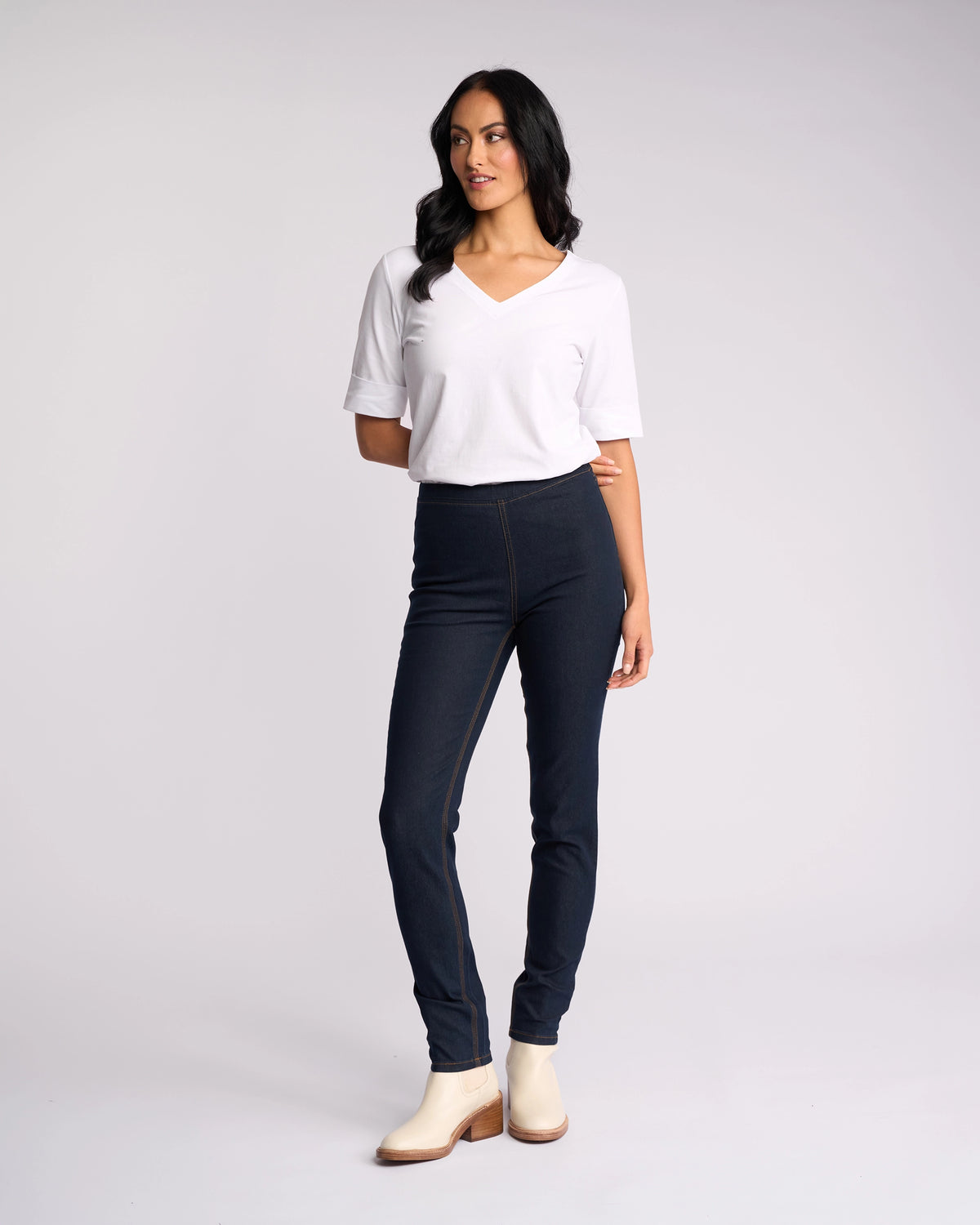 A woman with long dark hair wears a white V-neck top and Vassalli’s Origin Pull On - Indigo Denim (Contrast Stitch) skinny jeans with light ankle boots, standing against a white background and gazing slightly to the side.