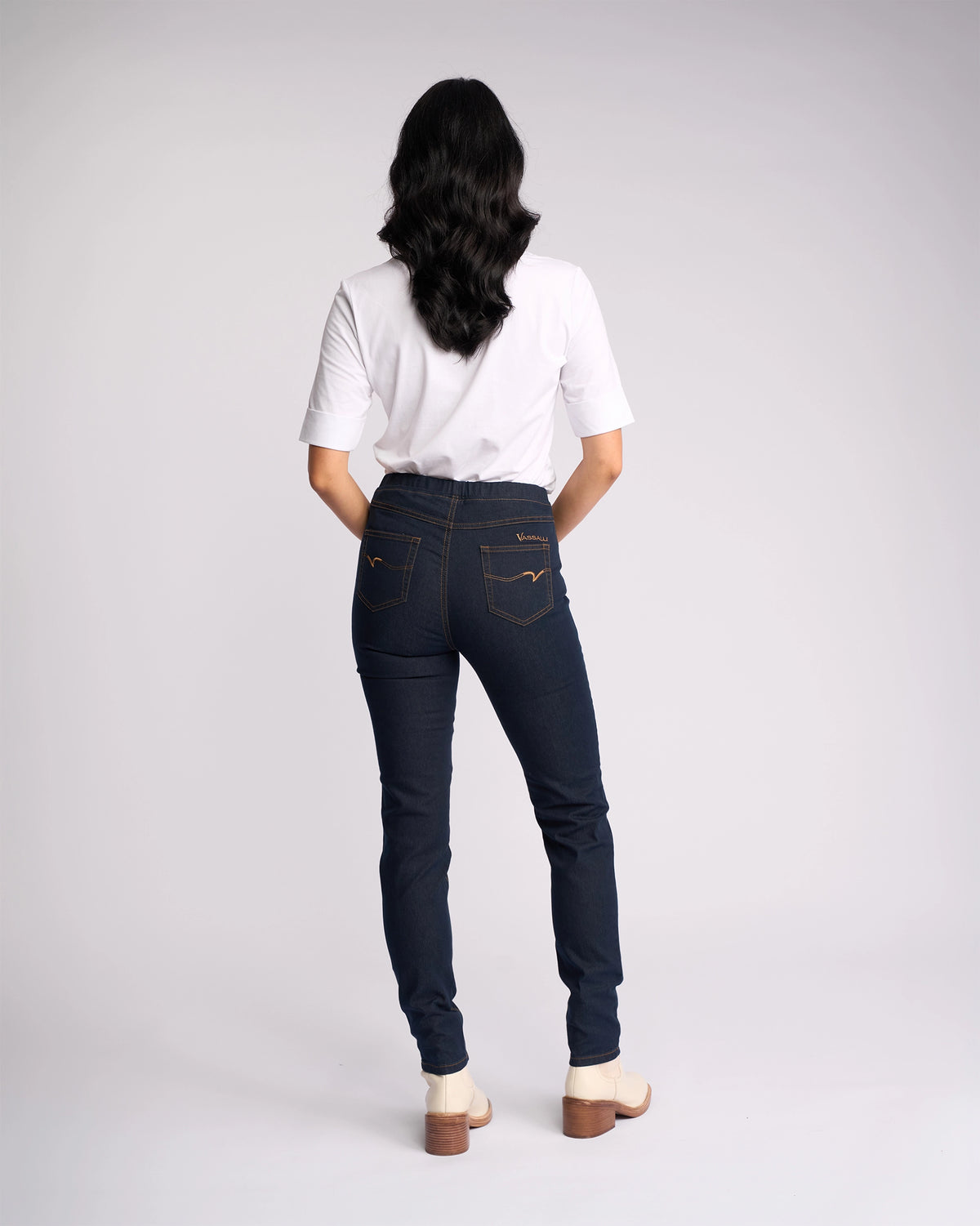 A person with long dark hair, in a white t-shirt and Vassalli Origin Pull On - Indigo Denim (Contrast Stitch) skinny-leg jeans, stands facing away from the camera against a plain light background.