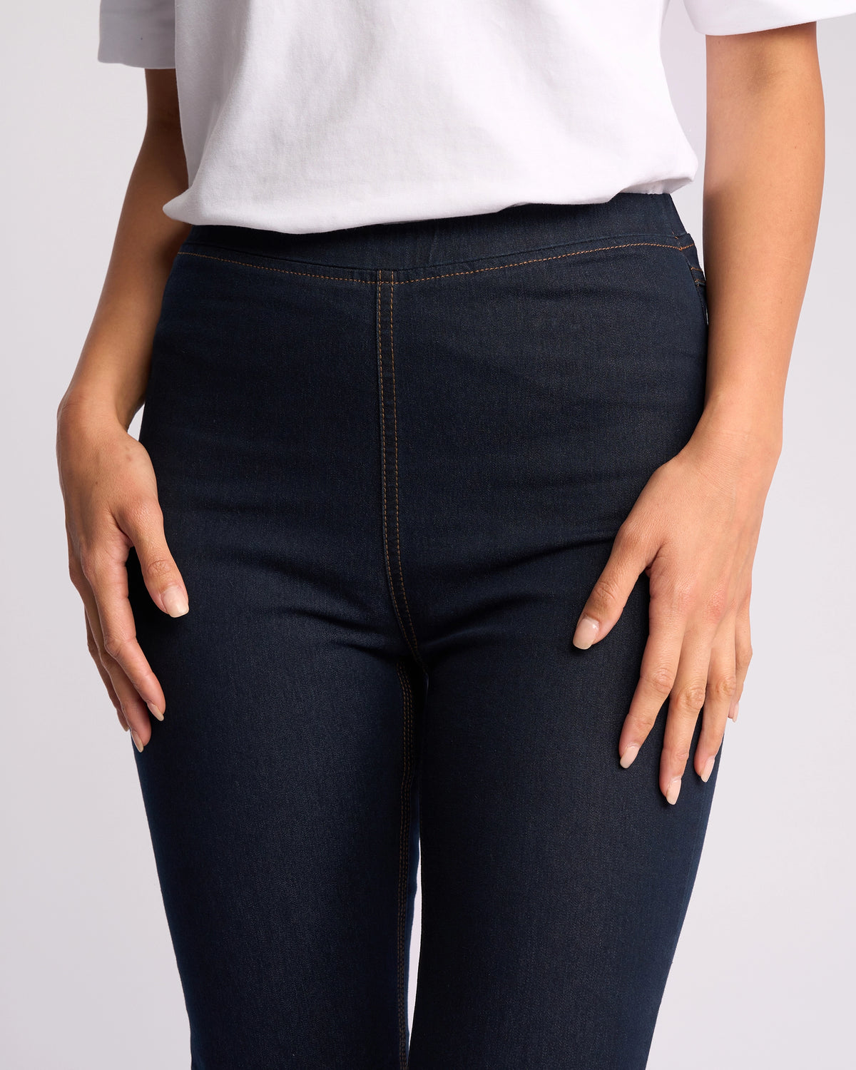 A person wears a white t-shirt and Vassalli Origin Pull On - Indigo Denim (Contrast Stitch) high-rise, skinny-leg jeans; only their torso and upper legs are visible with hands resting on thighs.