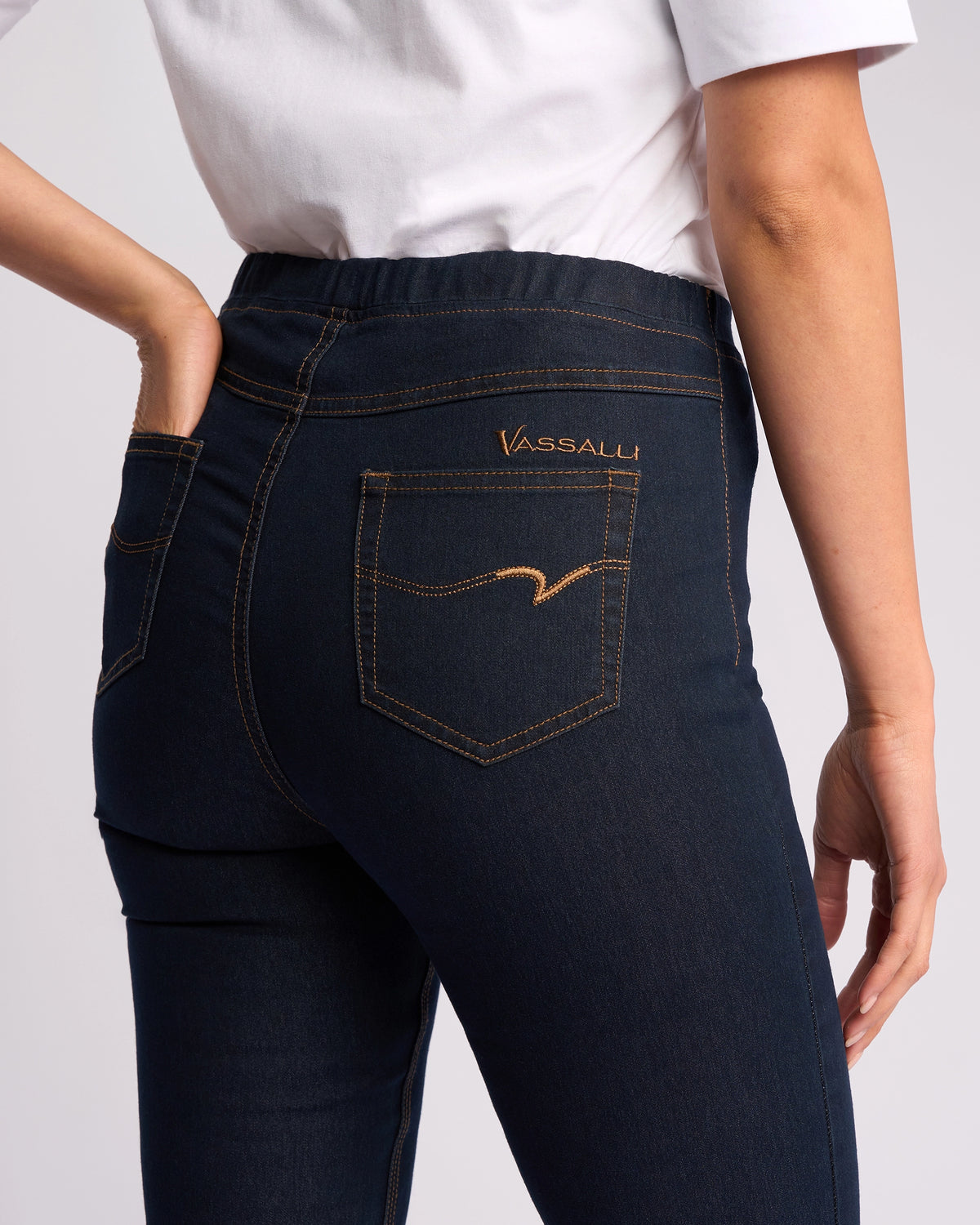 A person wearing Vassalli Origin Pull On - Indigo Denim (Contrast Stitch) jeans is shown from the waist down, paired with a white shirt, with one hand resting near the back pocket.