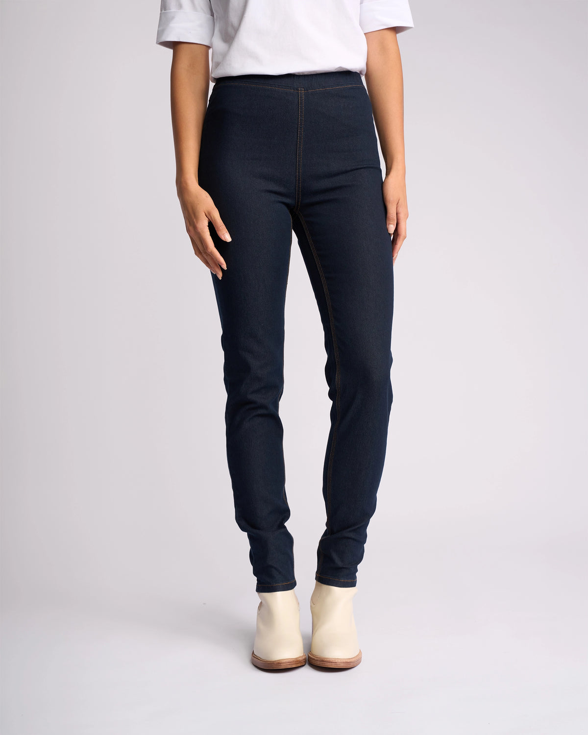 A person wears a white short-sleeve shirt, Vassalli Origin Pull On - Indigo Denim (Contrast Stitch) high rise skinny-leg jeans, and white ankle boots, standing against a plain light background with hands relaxed at their sides.