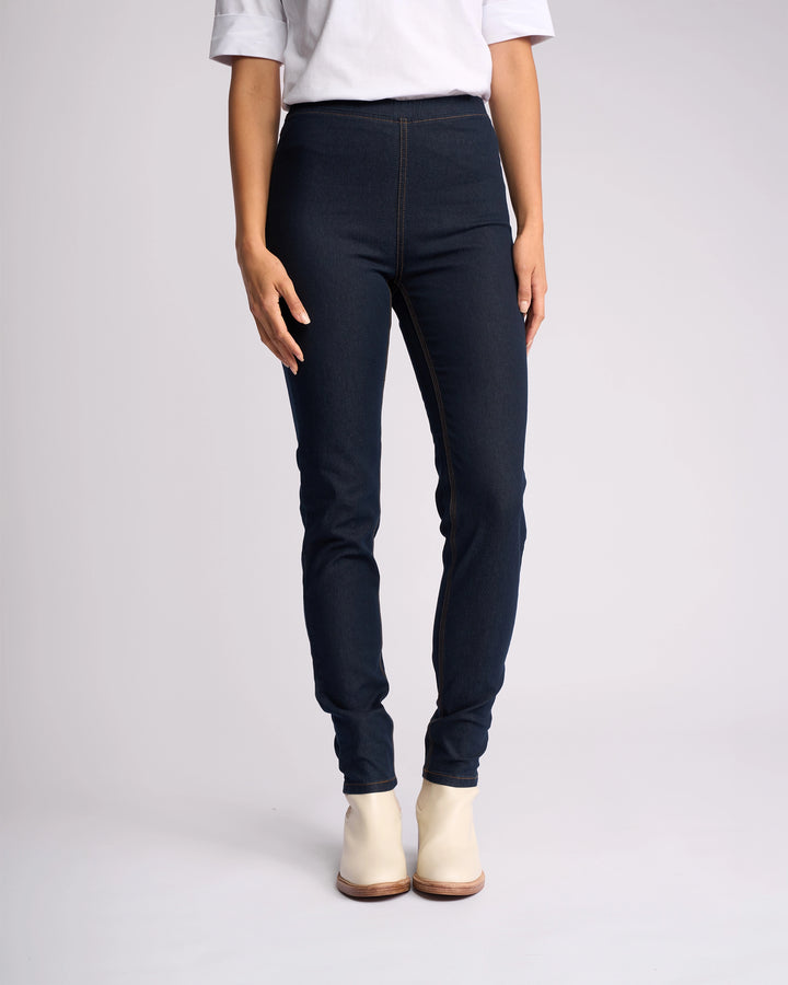 A person wears a white short-sleeve shirt, Vassalli Origin Pull On - Indigo Denim (Contrast Stitch) high rise skinny-leg jeans, and white ankle boots, standing against a plain light background with hands relaxed at their sides.