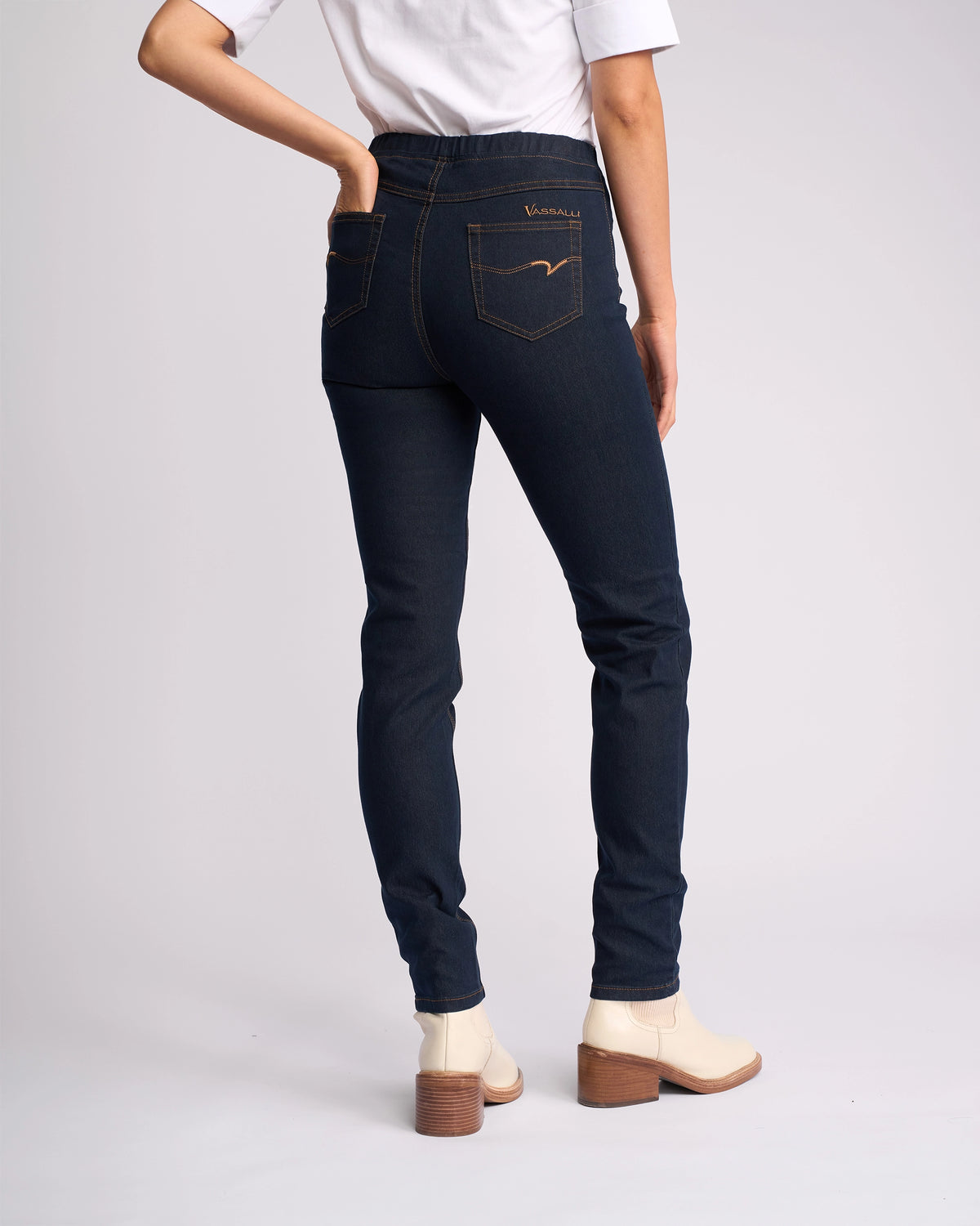 Wearing Vassalli's Origin Pull On - Indigo Denim (Contrast Stitch) jeans, paired with a white top and light ankle boots with a brown heel, a person stands facing away from the camera with one hand in their back pocket.